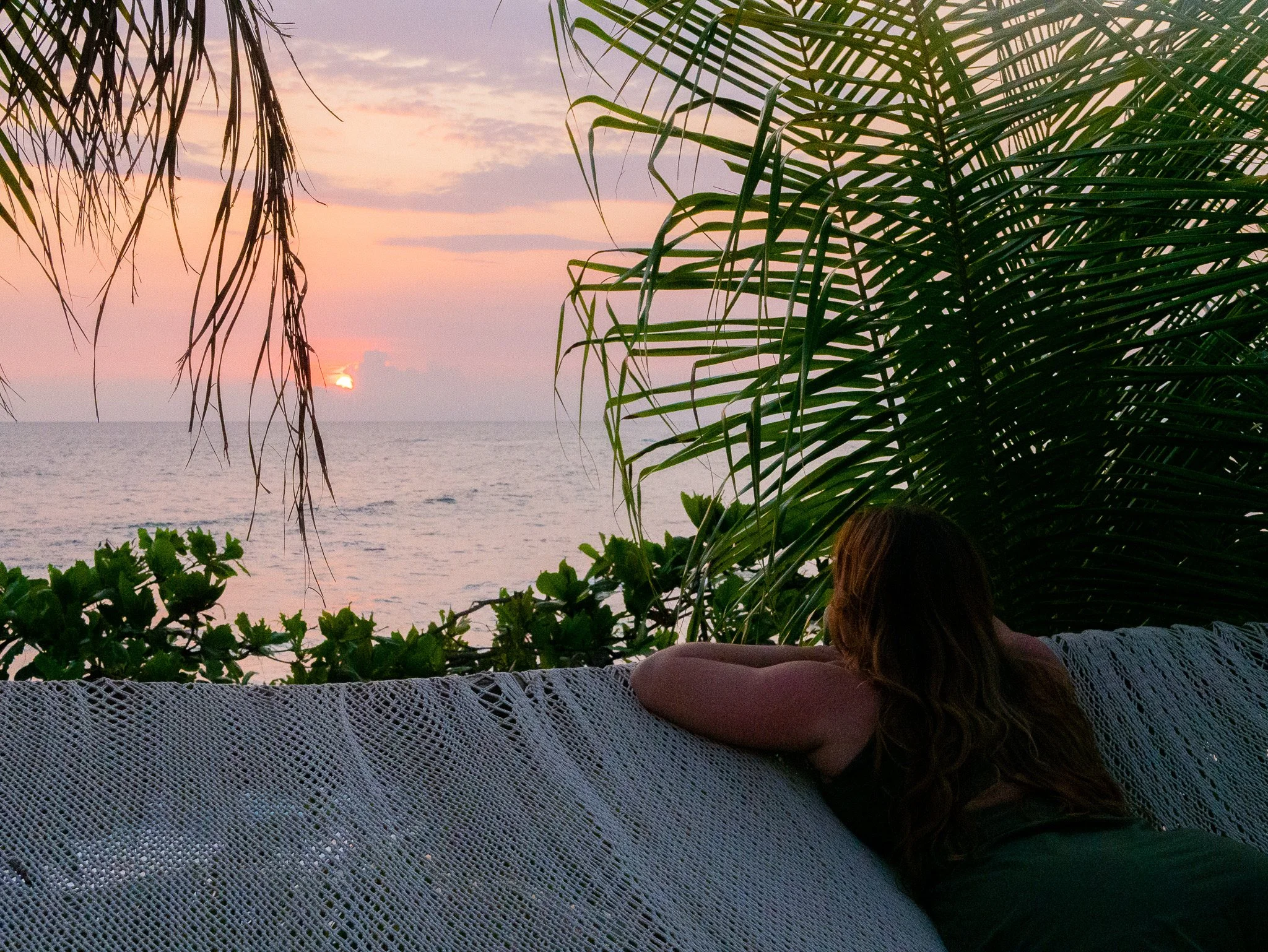 Woman resting on an oceanfront hammock watching a pink Pacific sunset through the palms at a Nicaragua retreat