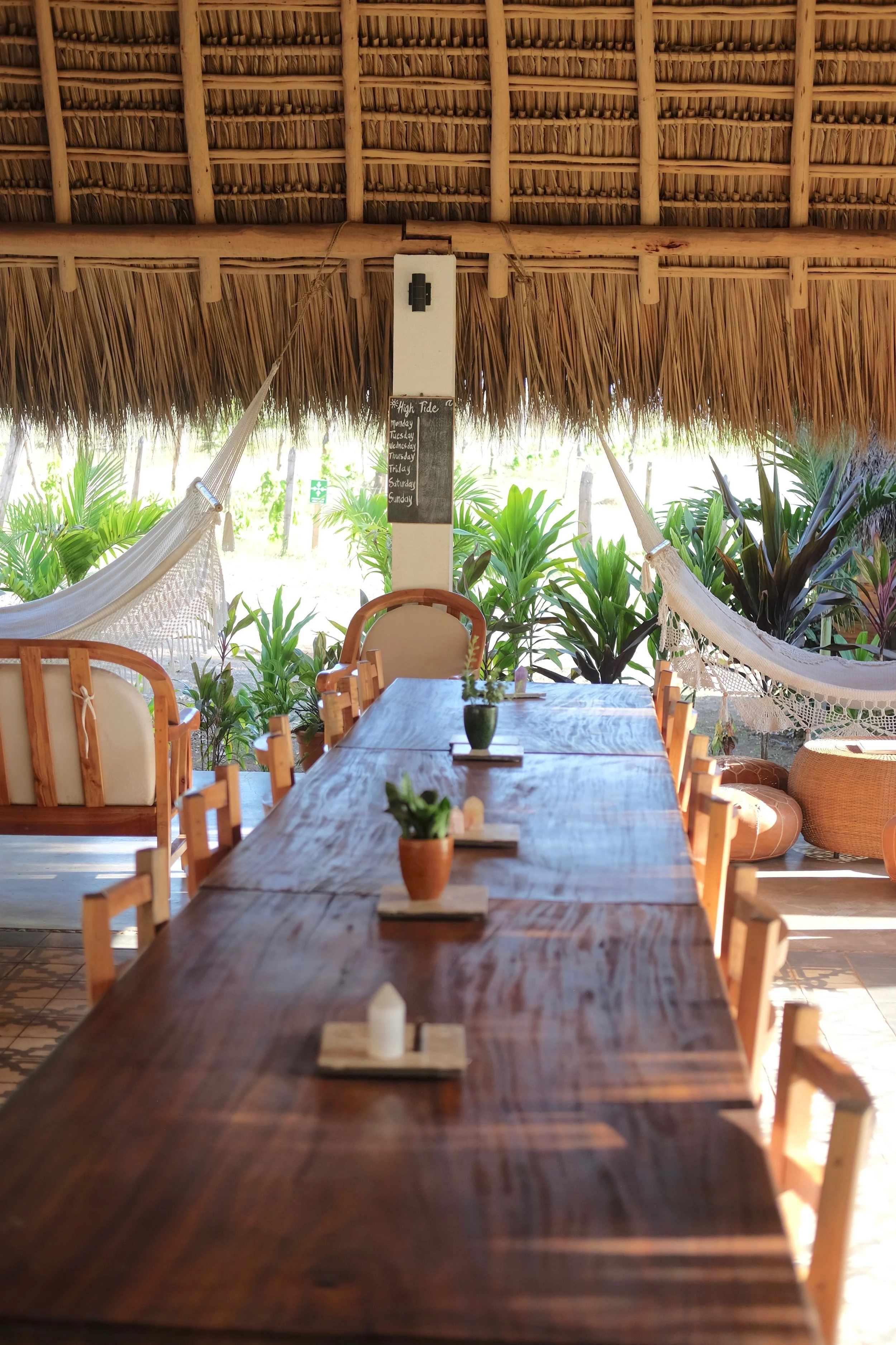 Long open-air wooden dining table under a thatched roof with hammocks and garden views at Still Salty Escape in Nicaragua