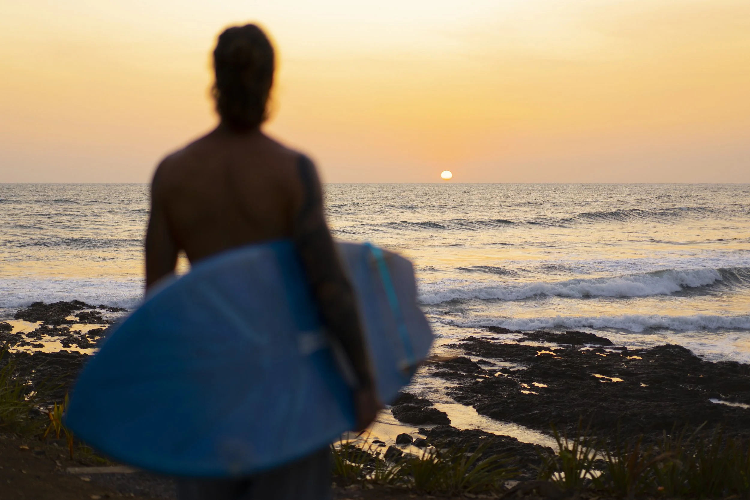Surfer holding a board watching the sun set into the Pacific from the rocks at a Nicaragua surf retreat