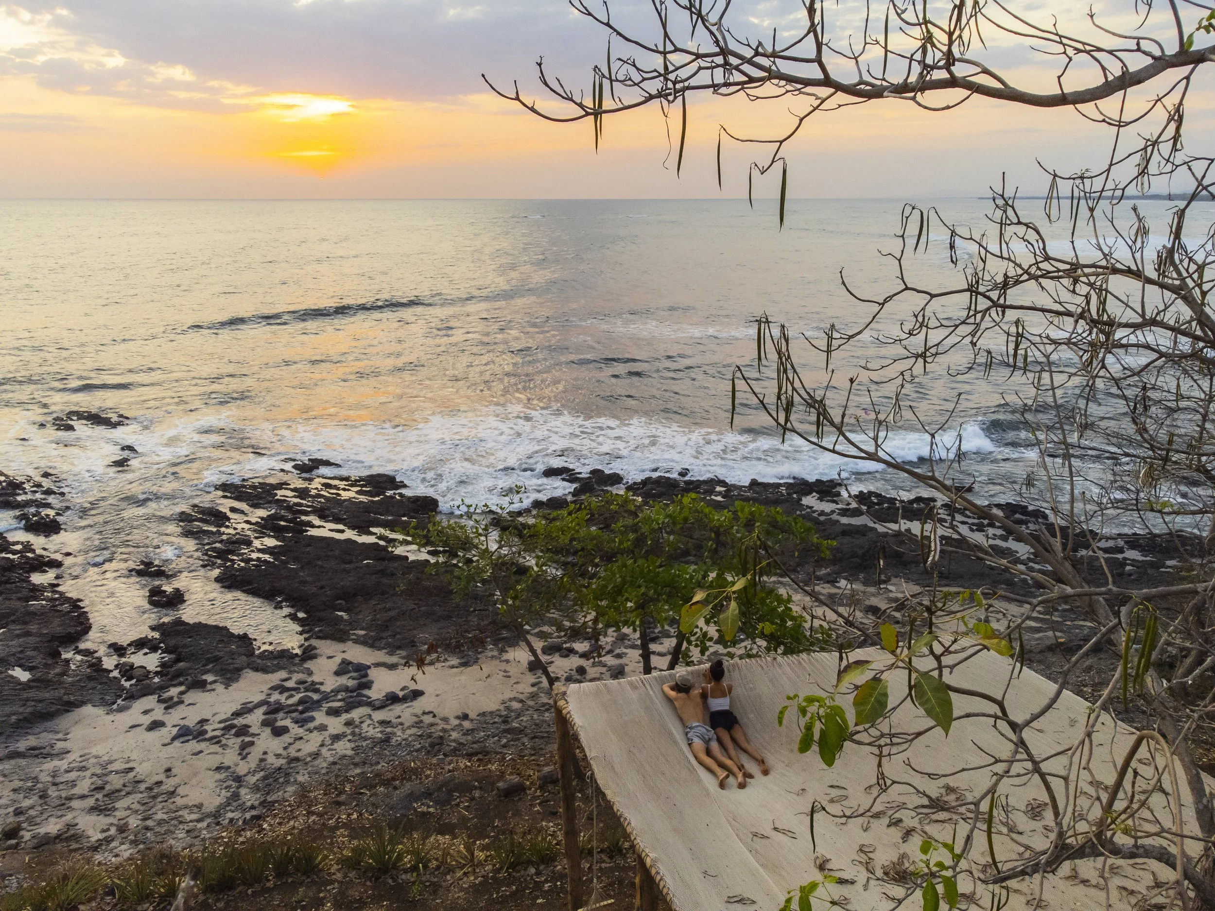 Aerial view of a couple relaxing on the giant net hammock above the rocky shoreline at sunset at Still Salty Escape Nicaragua