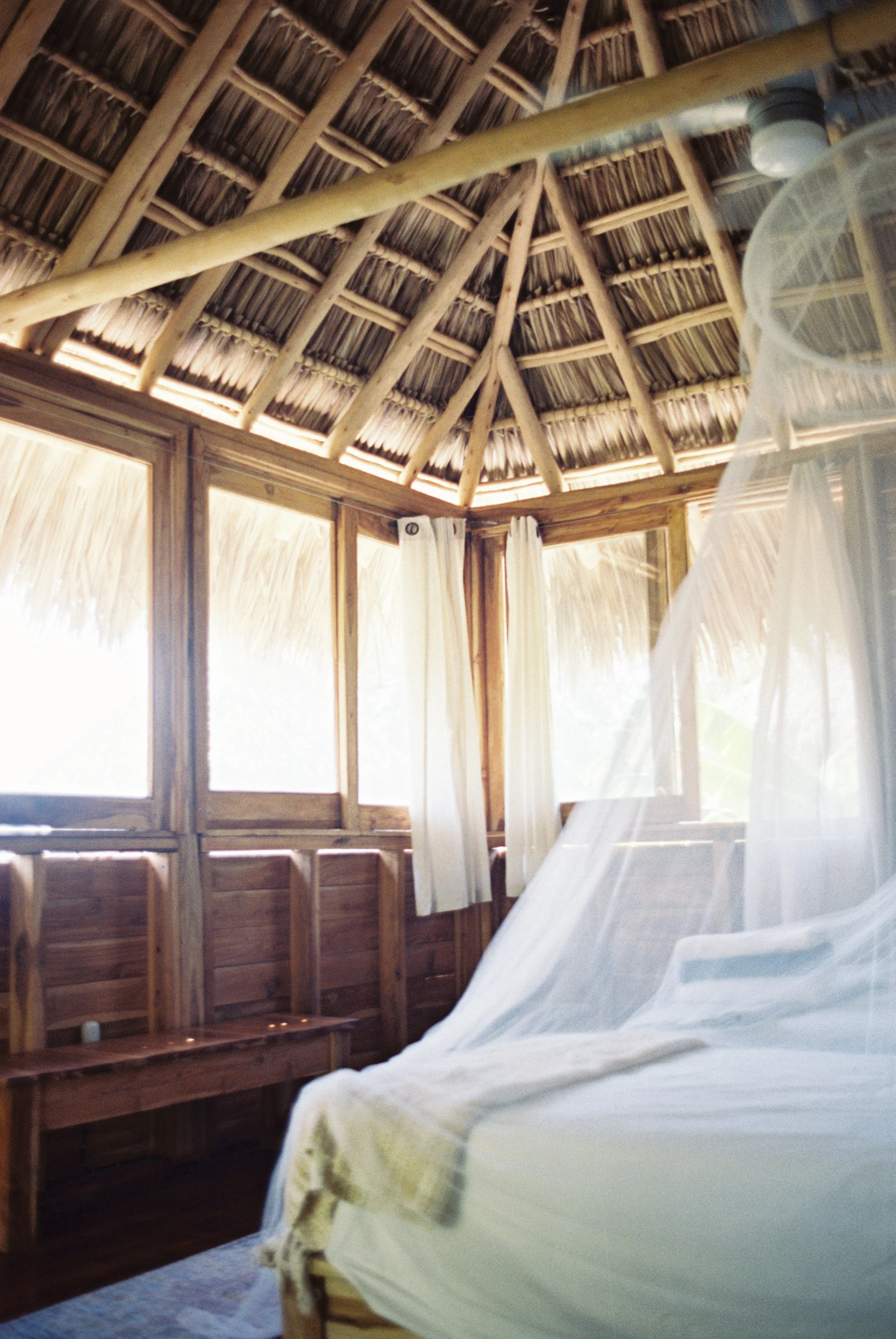 Upstairs double room with a mosquito-net bed, ceiling fan, and ocean-view windows under a conical thatched roof at Still Salty Escape Nicaragua