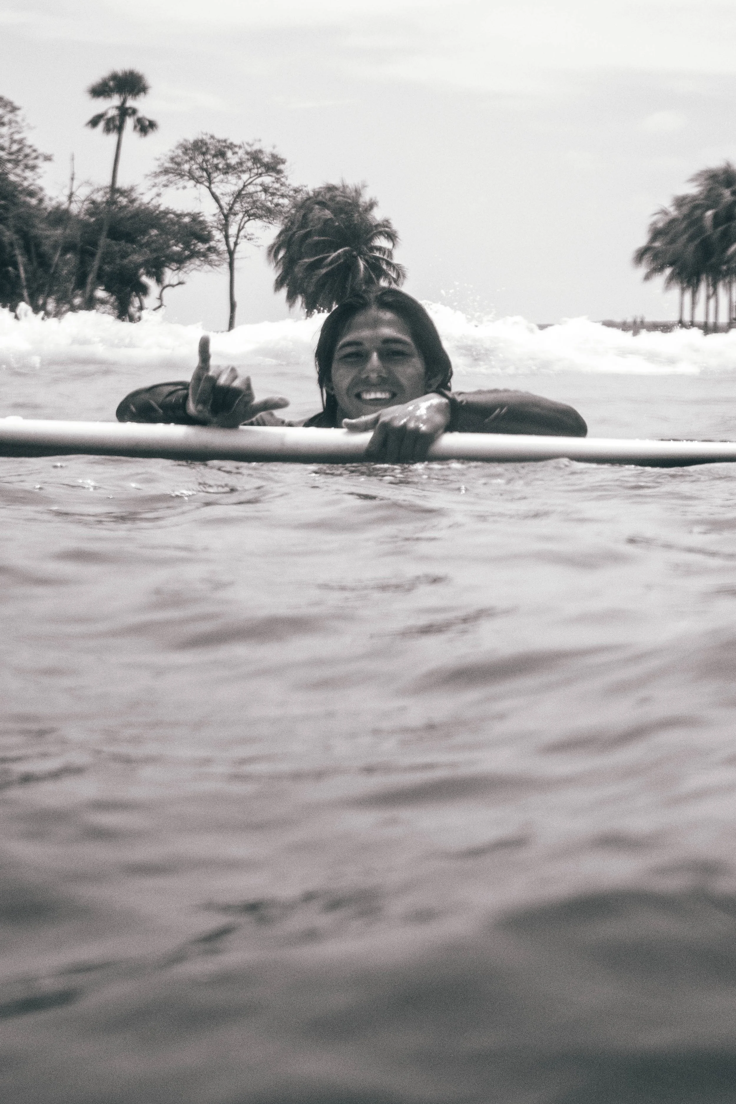 black and white photo of a surf instructor floating beside his board, smiling and making a shaka sign in an empty ocean lineup in Nicaragua