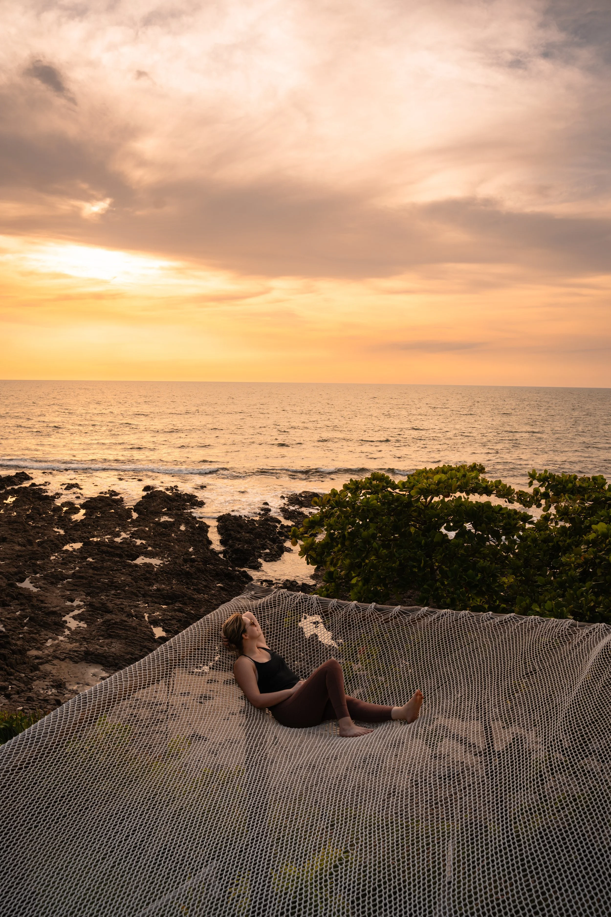 Guest lounging on the giant net hammock above the rocks at an orange Pacific sunset at a Nicaragua retreat