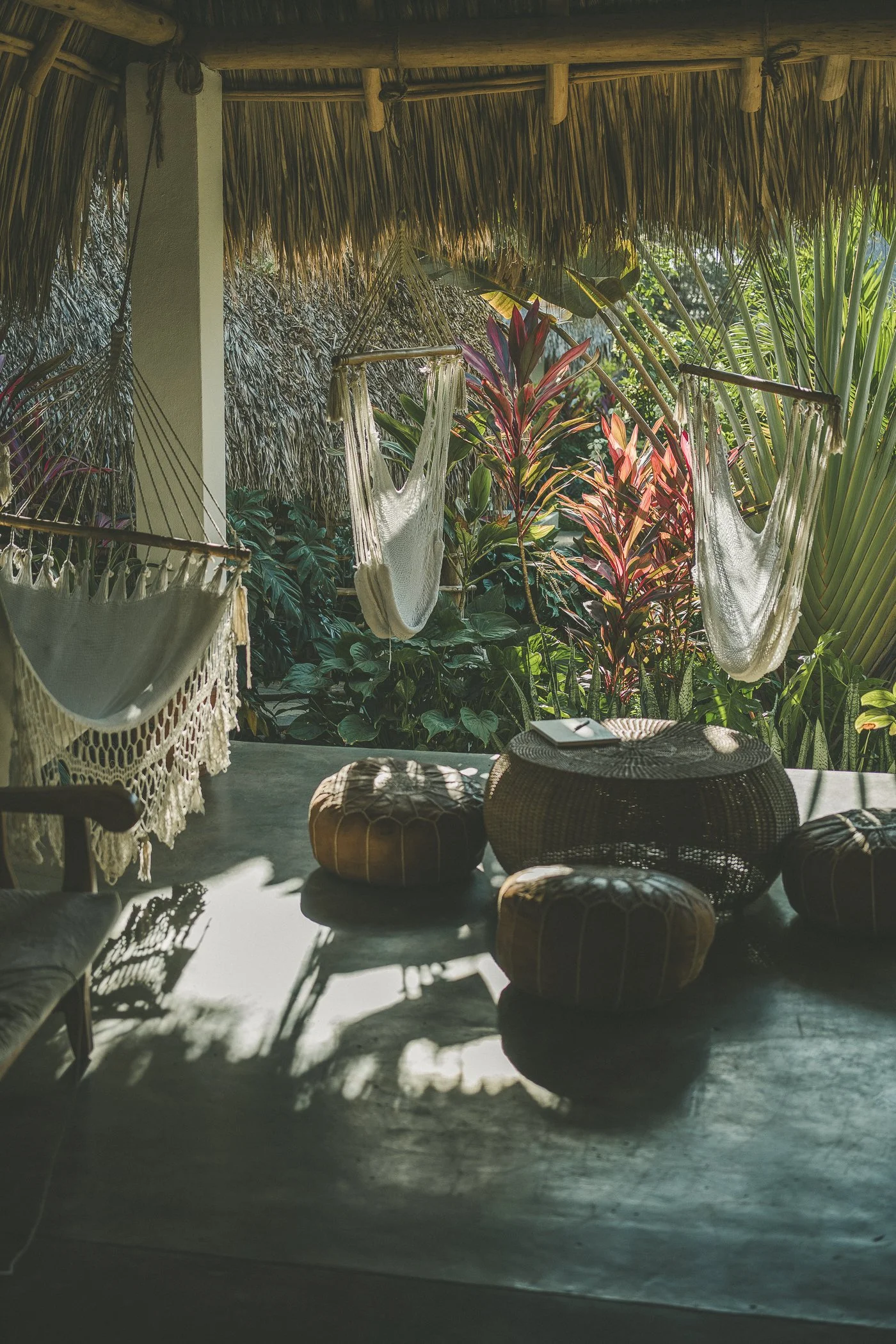 Covered open-air lounge with woven hammock chairs and leather poufs beside a tropical garden at a Nicaragua surf and yoga retreat