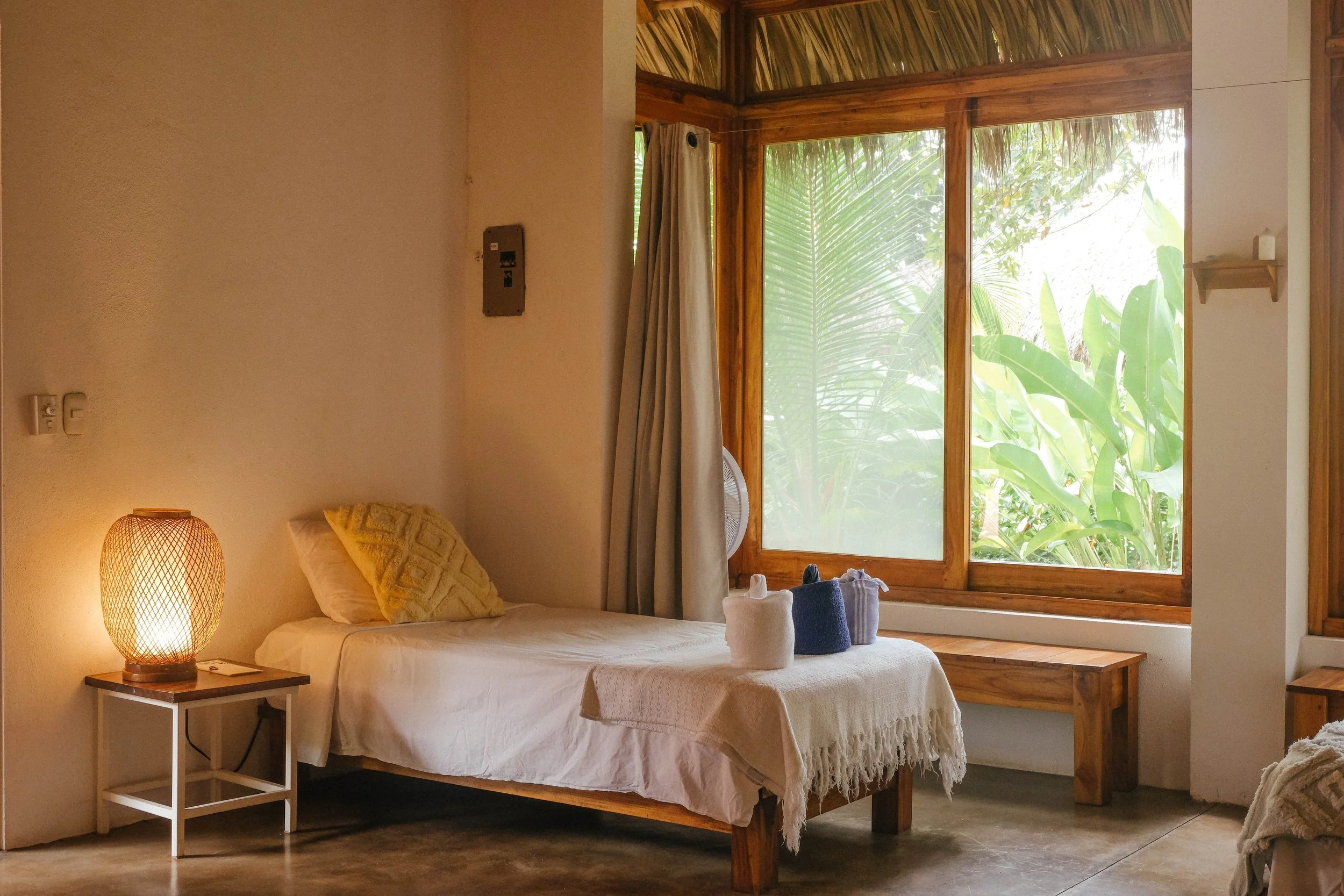 Single bed with a woven lamp beside a large garden-view window in an air-conditioned ensuite downstairs cabaña at Still Salty Escape Nicaragua