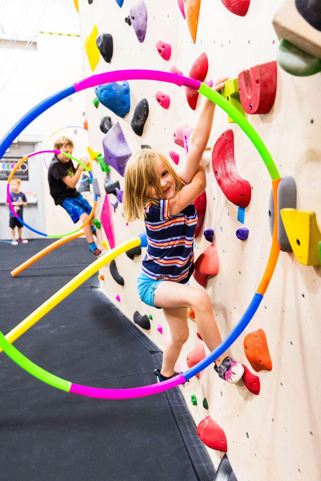 Children climbing on an indoor bouldering wall with colorful holds and hula hoops.