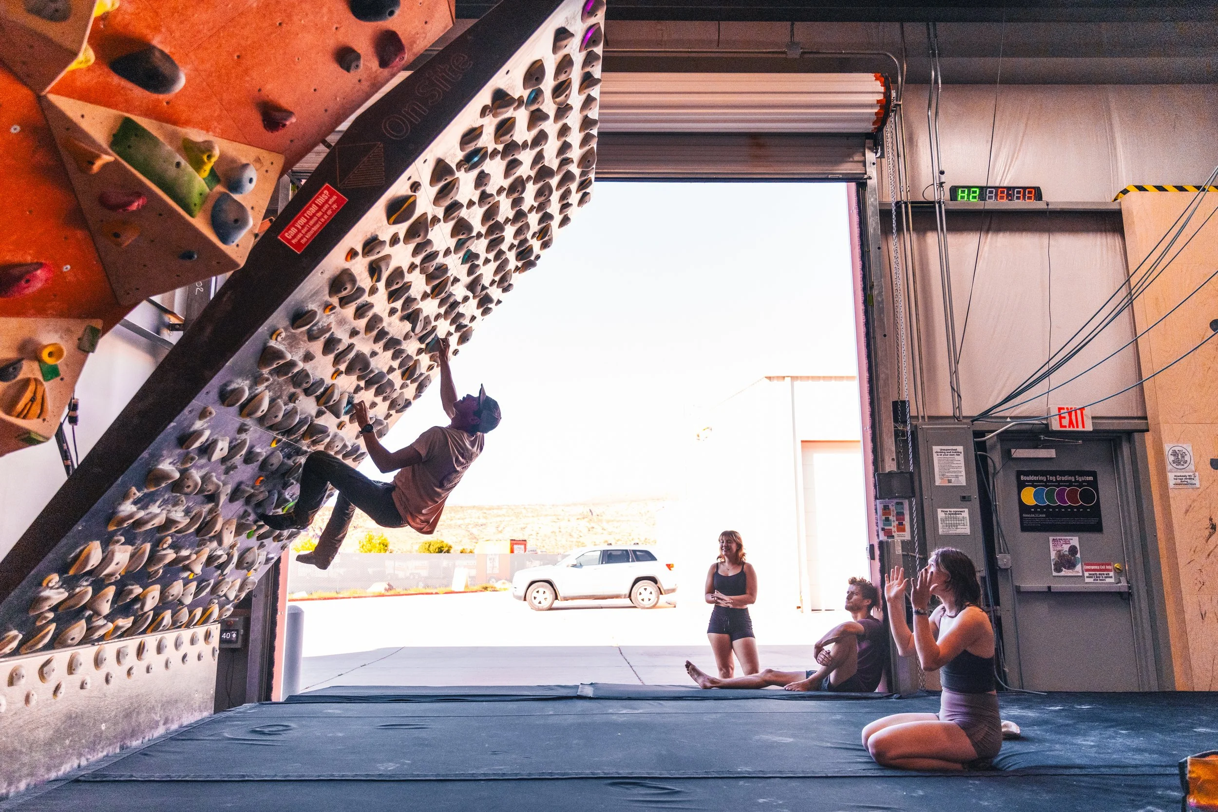 Indoor rock climbing gym with a climbing wall and people preparing for a climb. Some individuals are sitting on mats, chatting or observing, while one person is climbing the wall. The gym has a high ceiling and an open garage-style door leading outside, with a car visible outside.