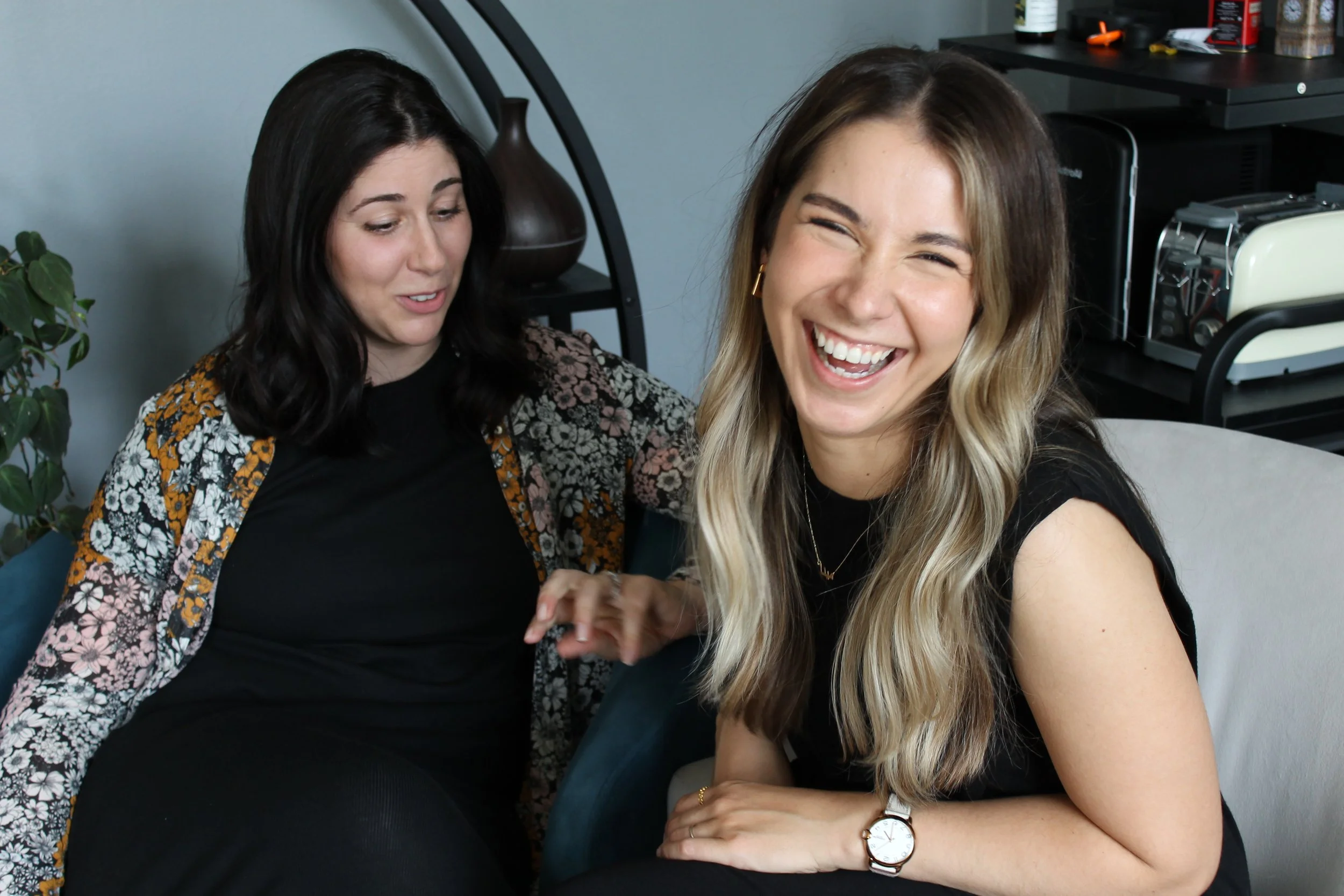 Two women sitting close together, smiling and laughing, in a casual indoor setting.