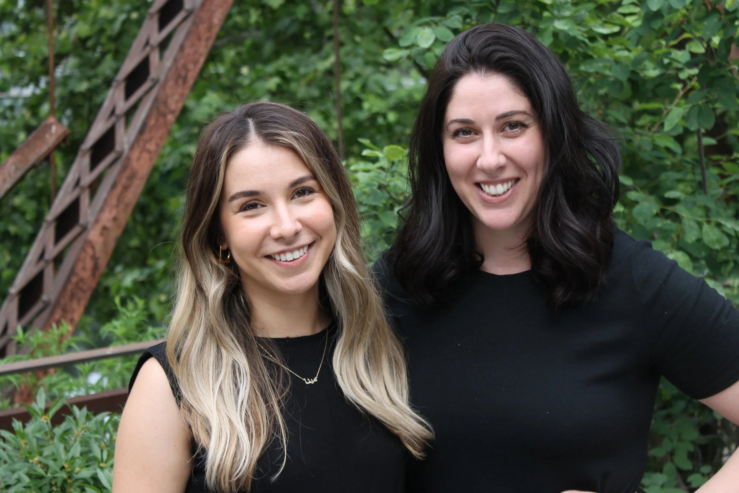Two women smiling outdoors, standing close together, with green foliage and a wooden structure in the background.