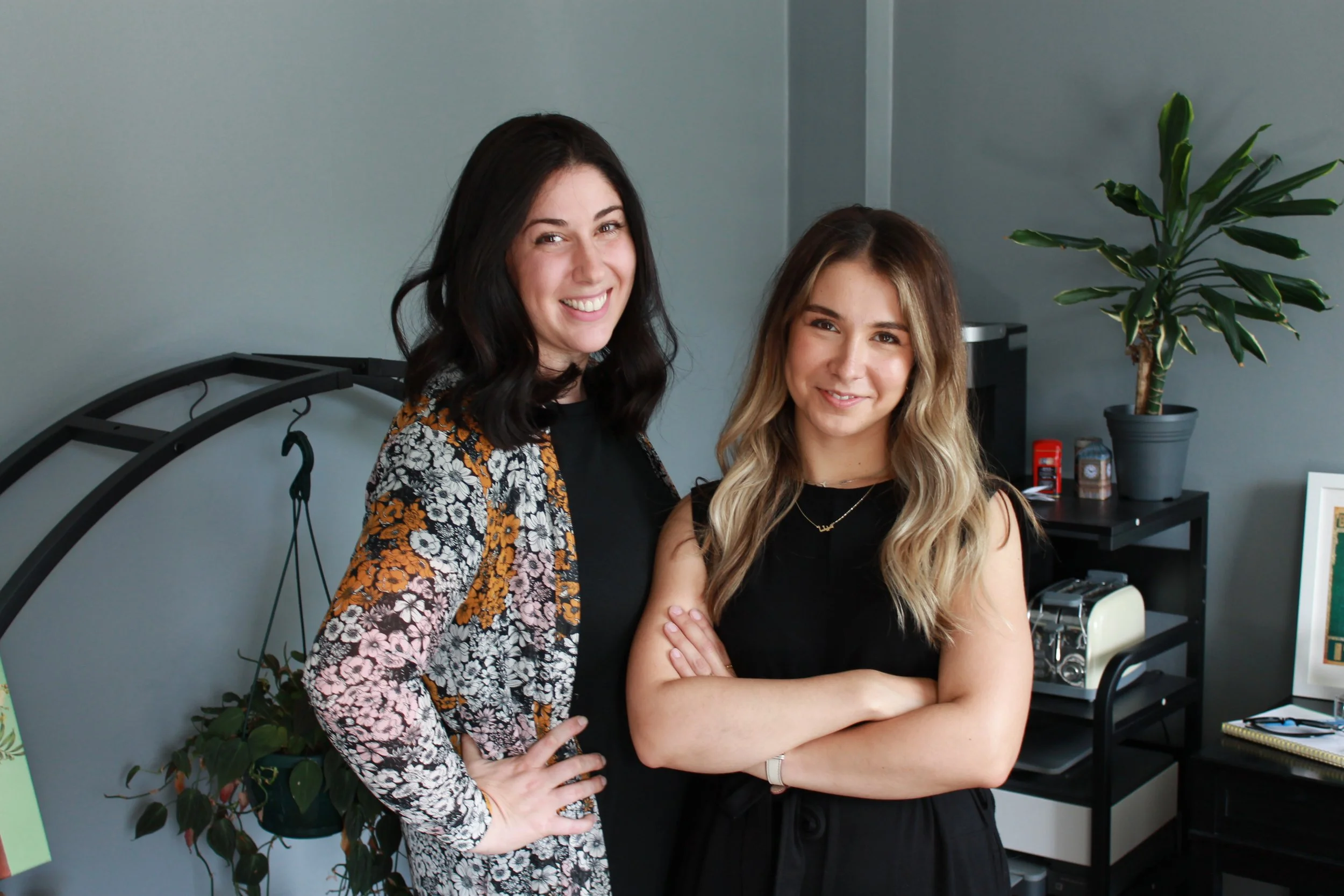 Two women standing indoors, smiling at the camera, with a plant and office supplies in the background.
