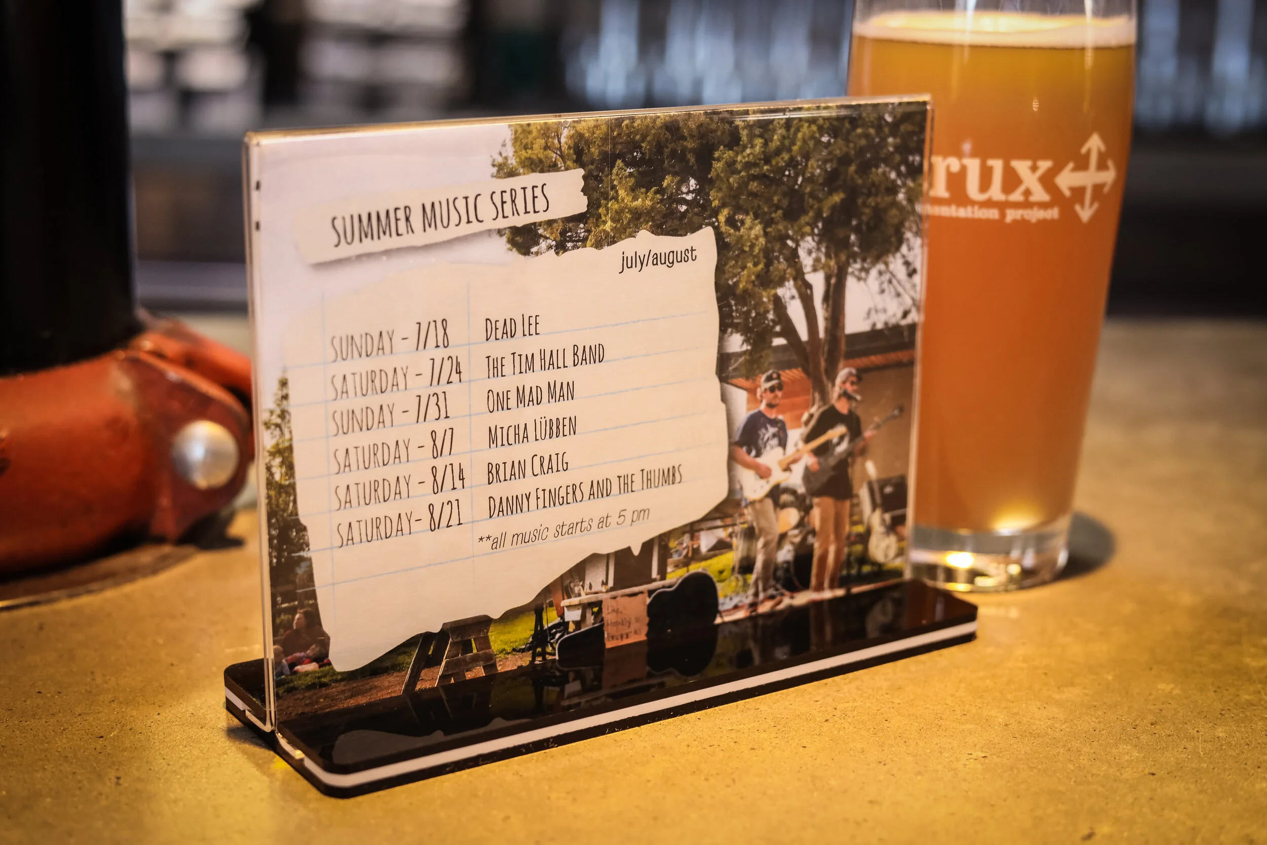 A music event schedule on a small stand with dates and bands, placed on a yellow table next to a glass of beer.