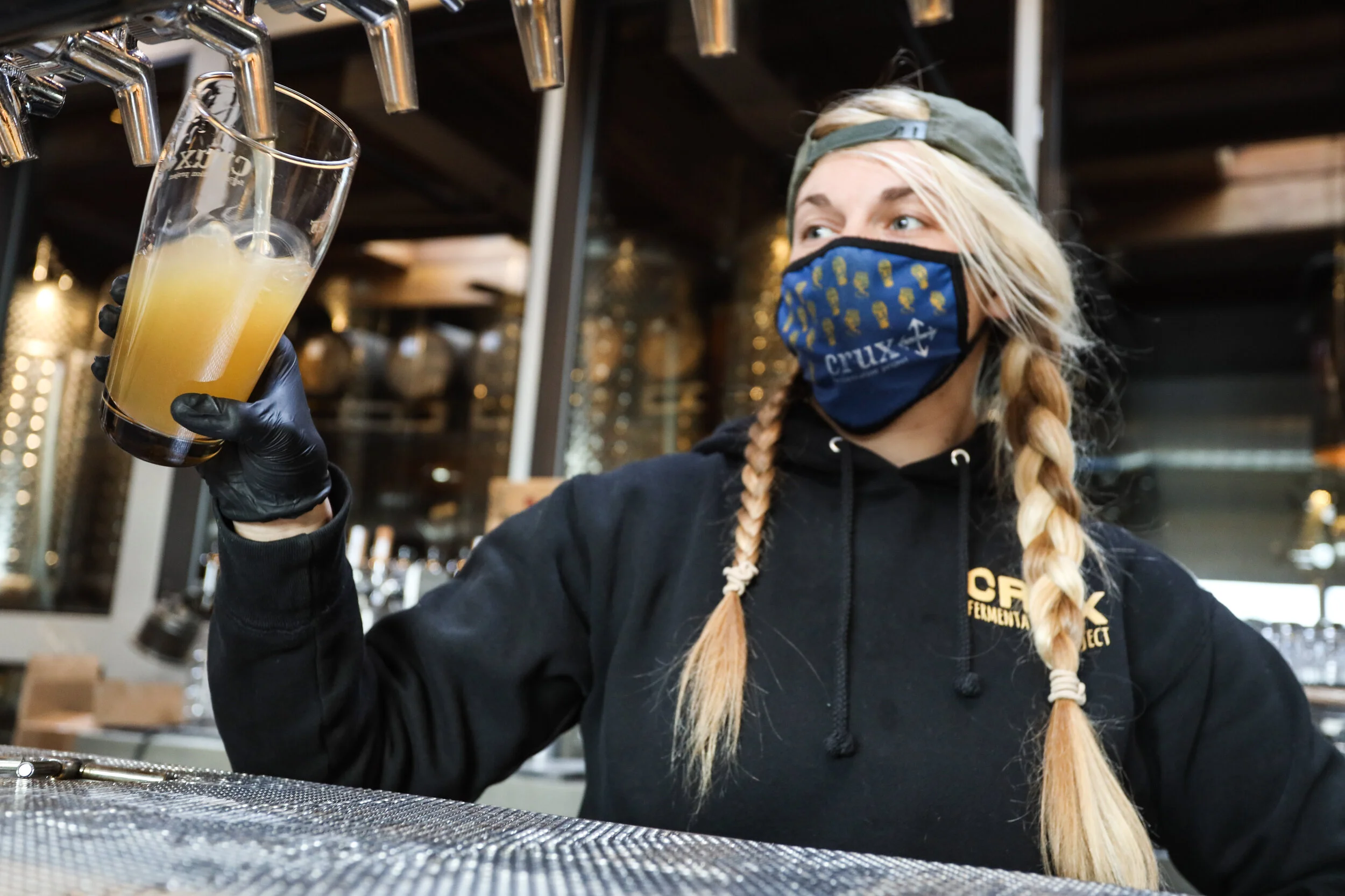 A woman wearing a black hoodie, a blue face mask, and a cap, holding a glass of light-colored beverage at a bar or brewery.