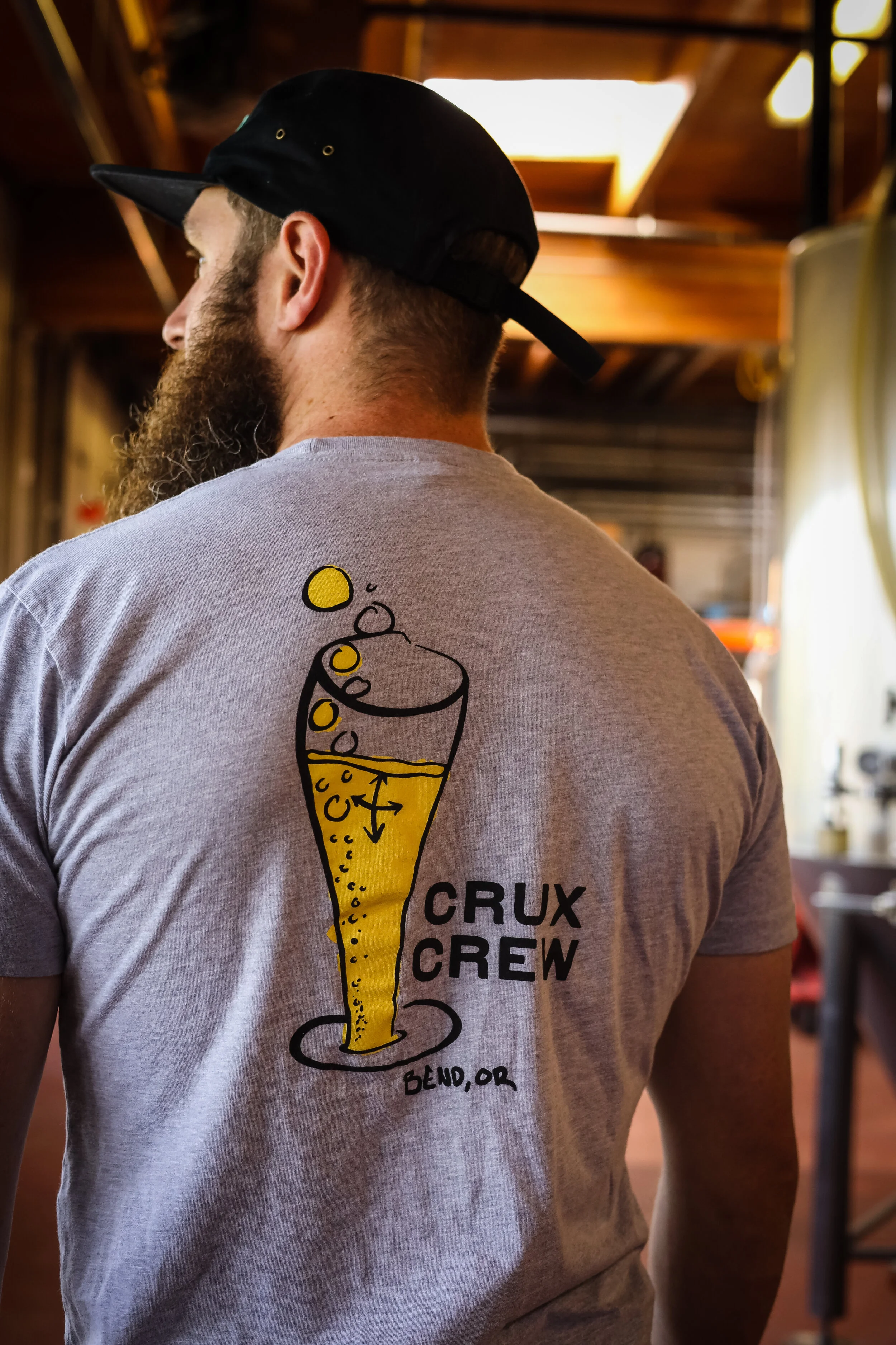 Person wearing a gray T-shirt with a graphic of a beer glass and the words "Crux Crew Bend, OR," in a cozy indoor setting.