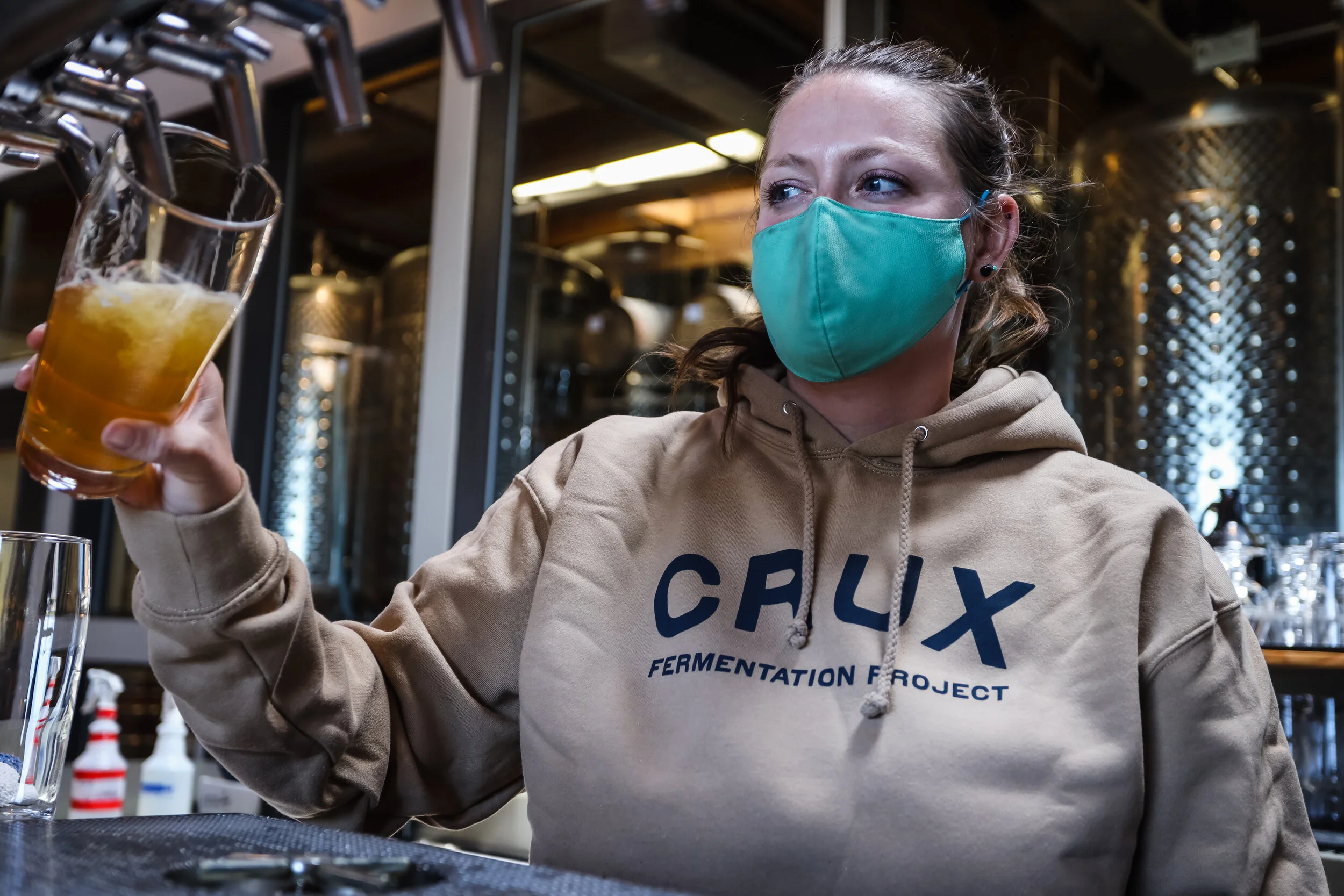 A woman wearing a teal face mask and a beige hoodie with the words 'CRUX FERMENTATION PROJECT' printed on it, holding a pint glass of beer in a bar or brewery setting.
