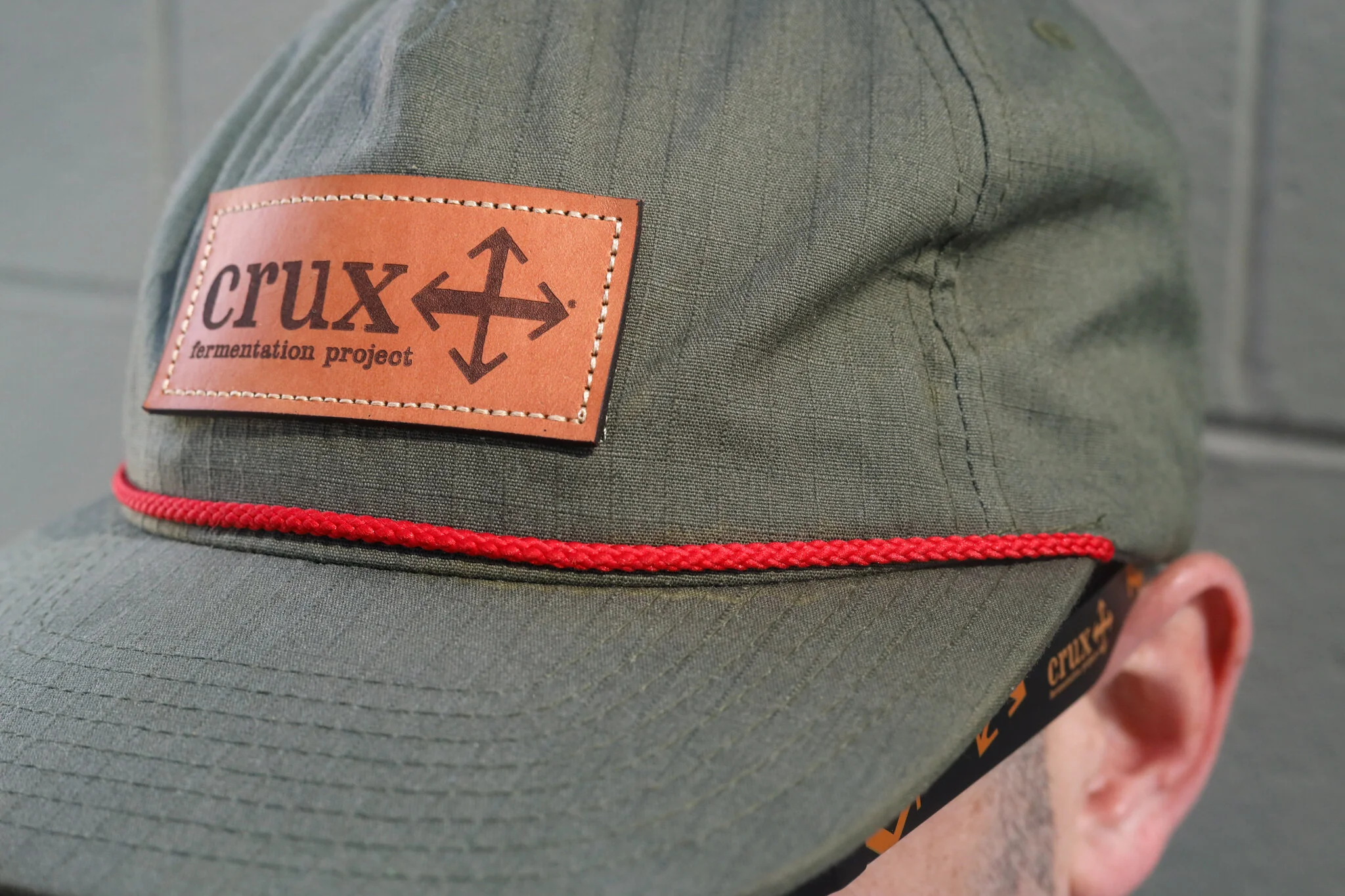 A green military-style cap with a brown leather patch that reads 'crux fermentation project' and a black arrow symbol. The cap features a red rope band above the brim, and part of an ear and neck are visible.