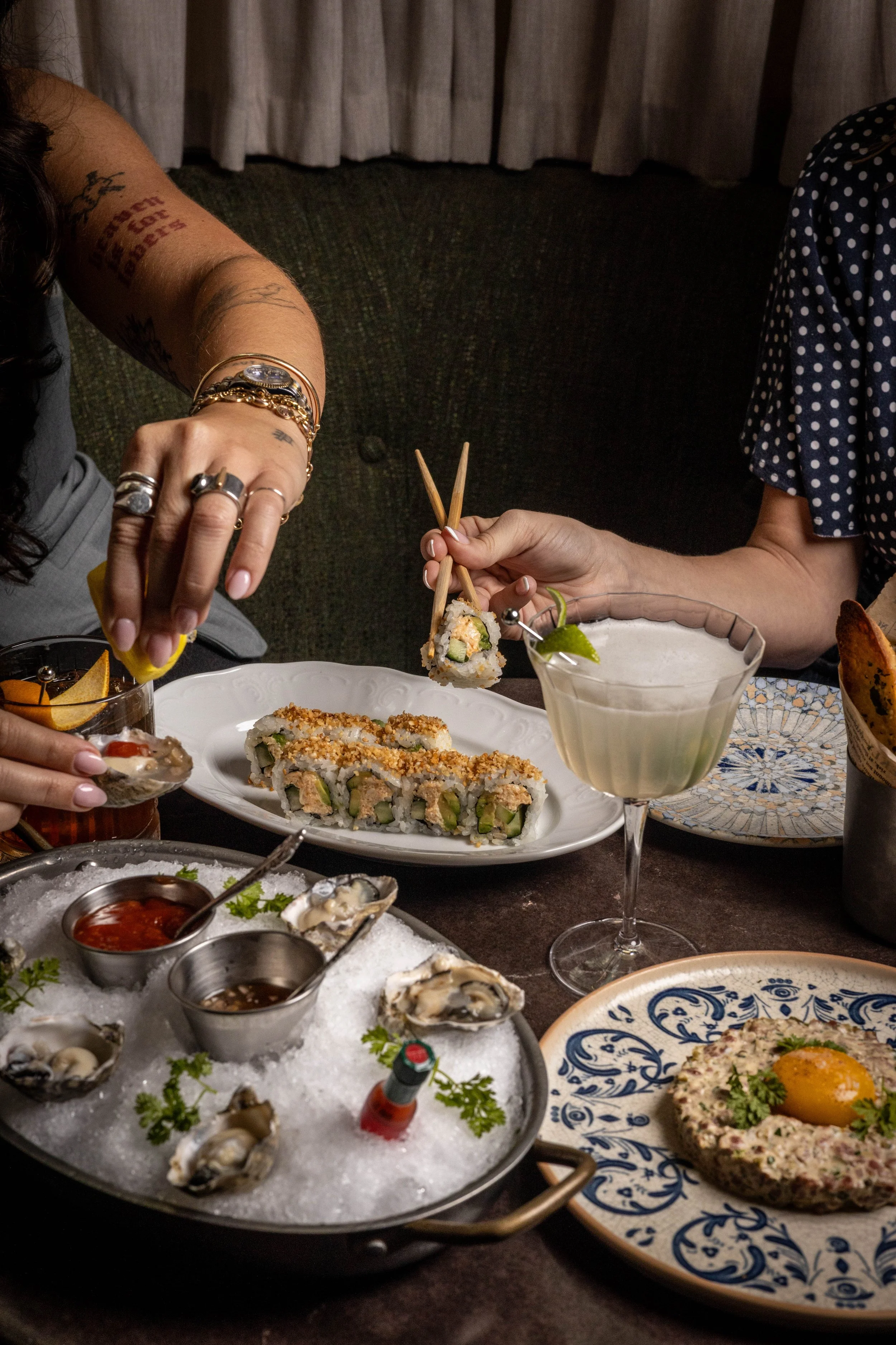 Two people sharing a meal at a table with sushi, oysters, and cocktails.