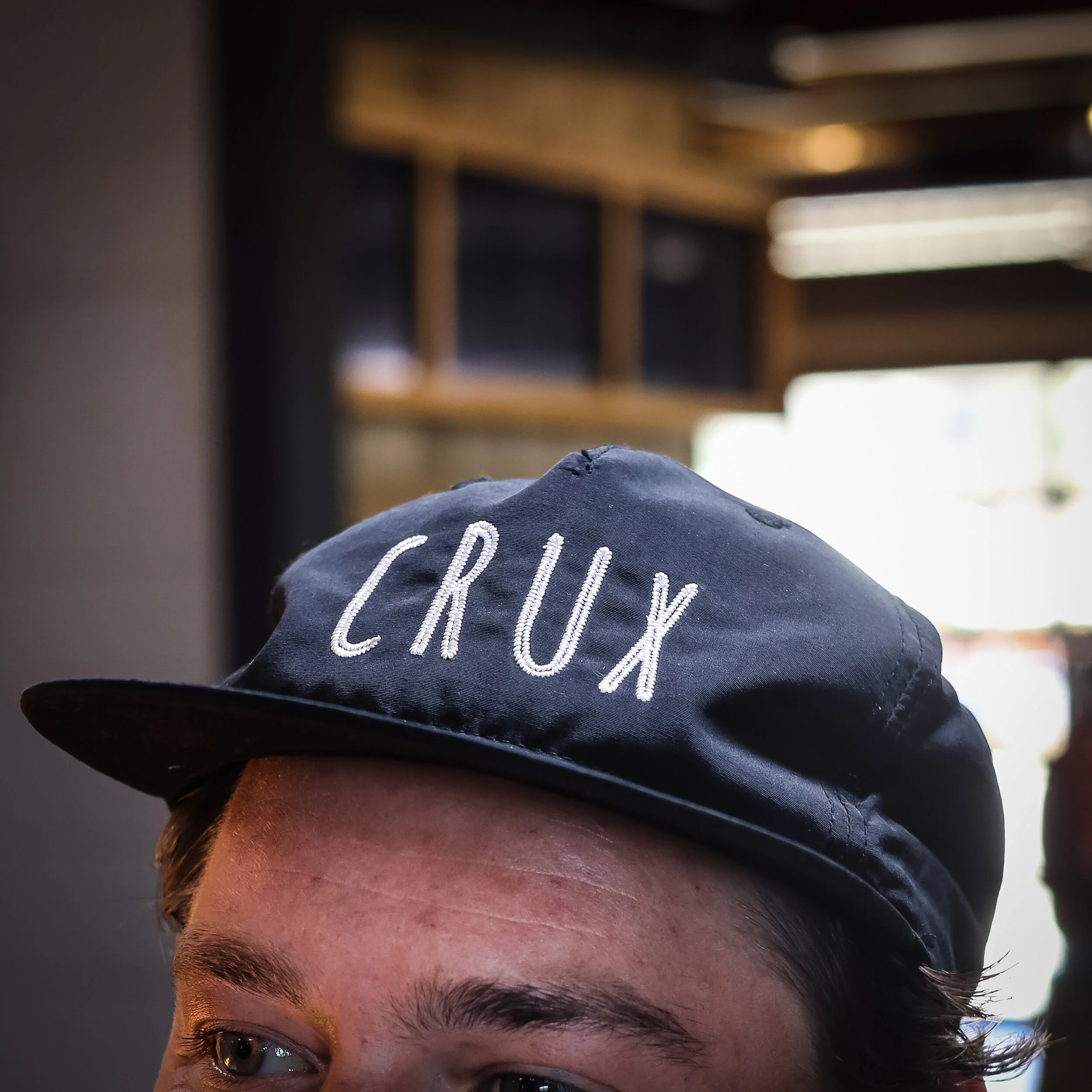 Close-up of a person's head wearing a dark baseball cap with 'CRUX' embroidered on it, showing part of their forehead, eyes, and eyebrows, in an indoor setting with a blurred background.
