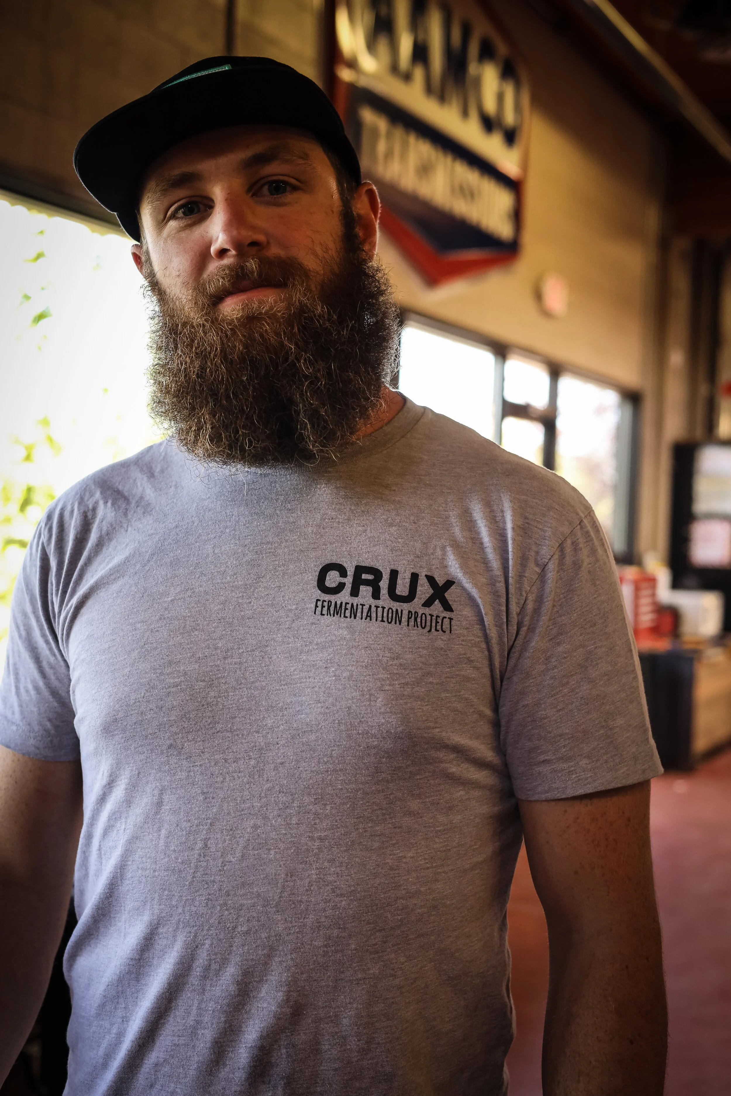 A man with a beard wearing a gray T-shirt that says 'CRUX Fermentation Project' and a black baseball cap, standing indoors with a sign and windows in the background.