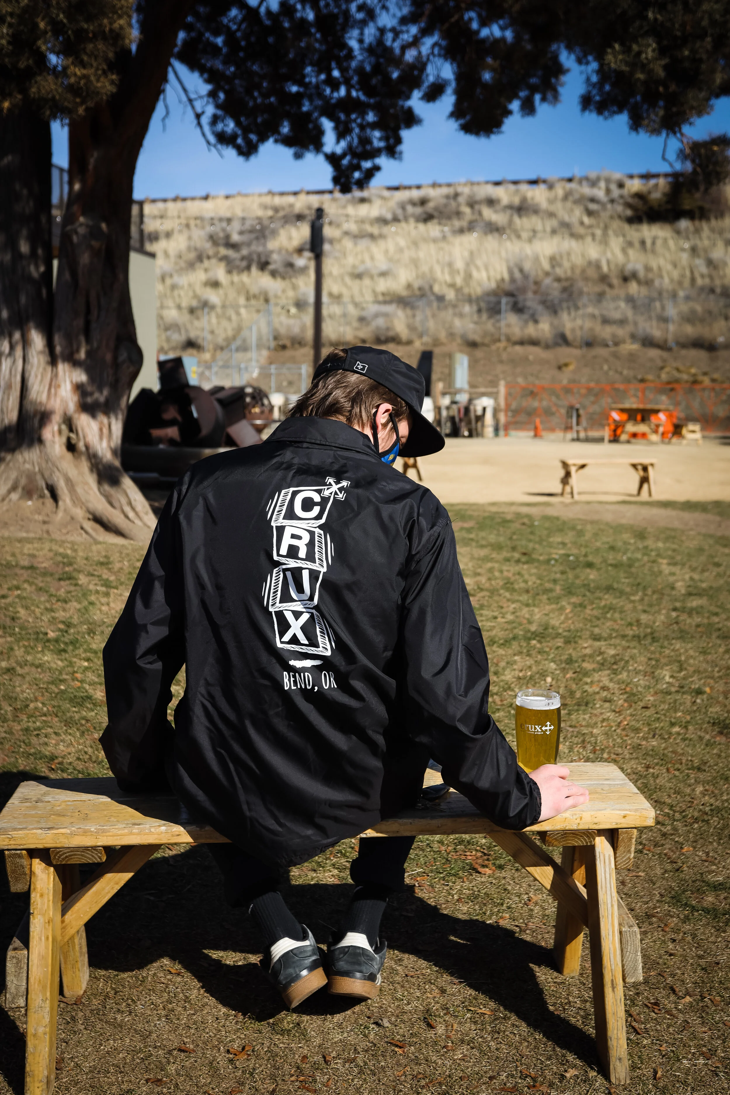 A person sitting on a wooden bench outdoors in a park, wearing a black jacket with 'C R U X Bend, OR' on the back, a black cap, face mask, and black shoes with white socks, holding a glass of beer.