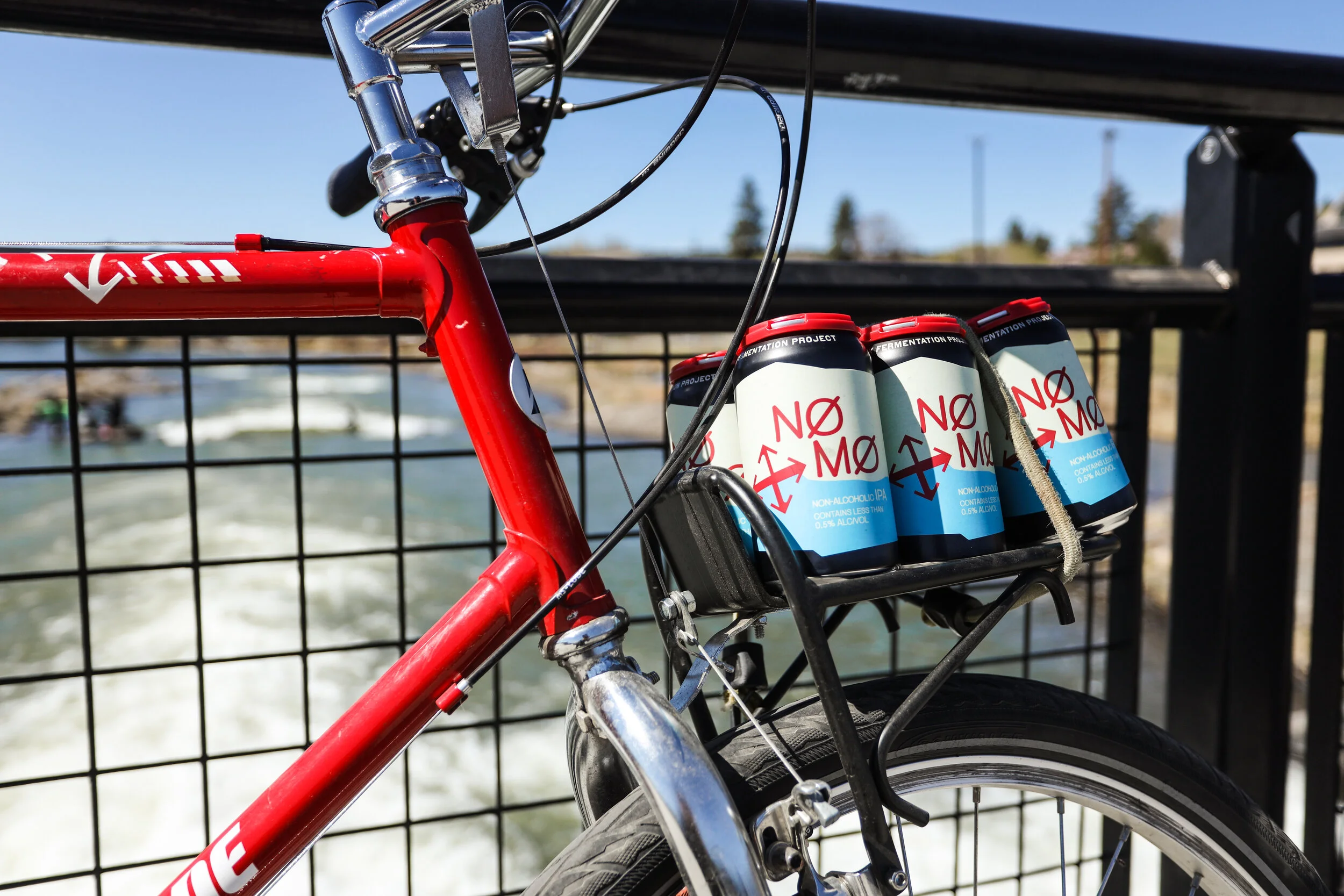 A red bicycle with a rear rack holding four cans of NO MØ non-alcoholic IPA, parked on a bridge with a river in the background.