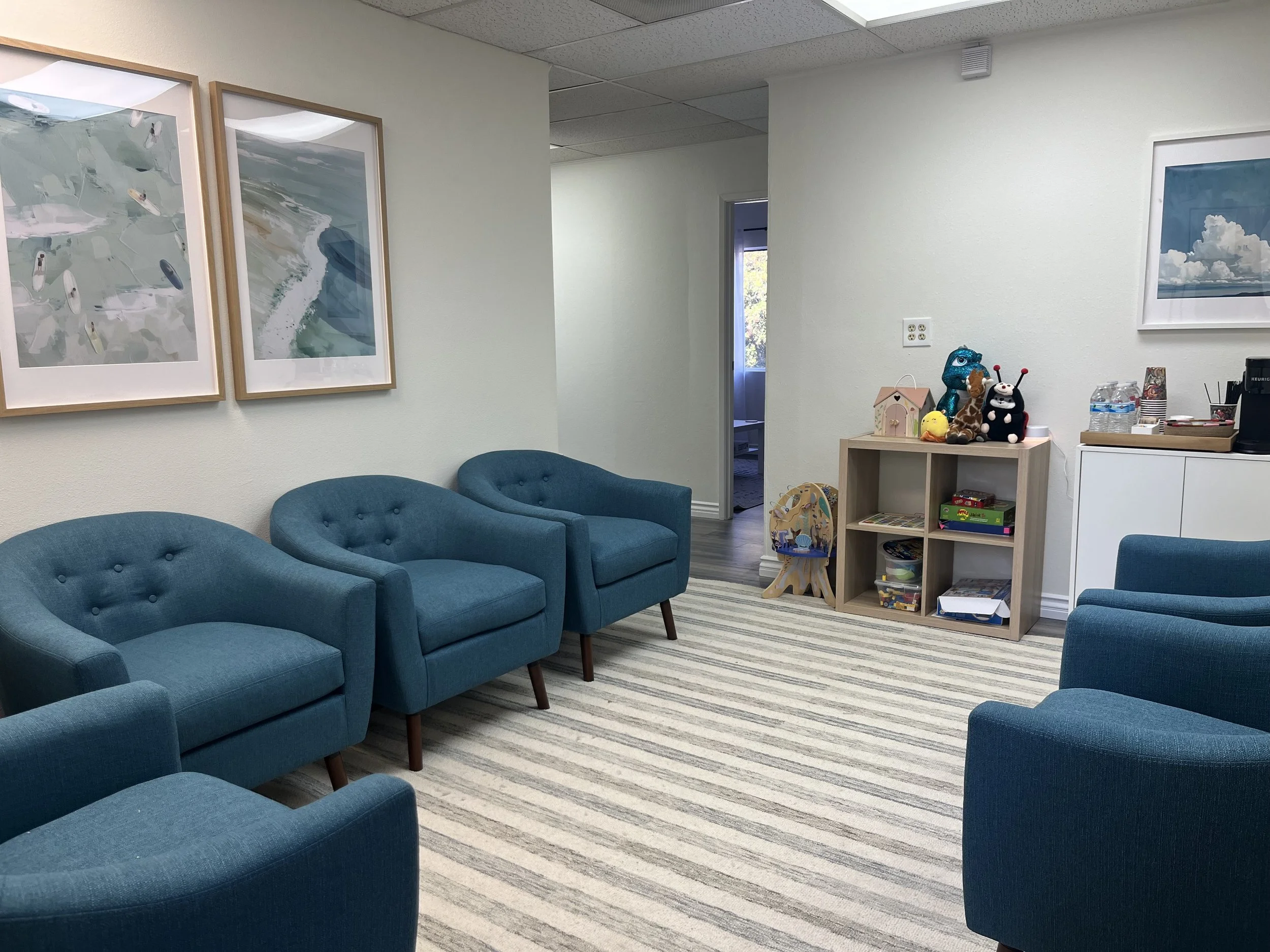 Waiting room of Carlsbad's Child Psychology Center. Blue, size-inclusive, overstuffed chairs are around the room and on top of a stripped rug. Cubed cabinet with toys is against the far wall