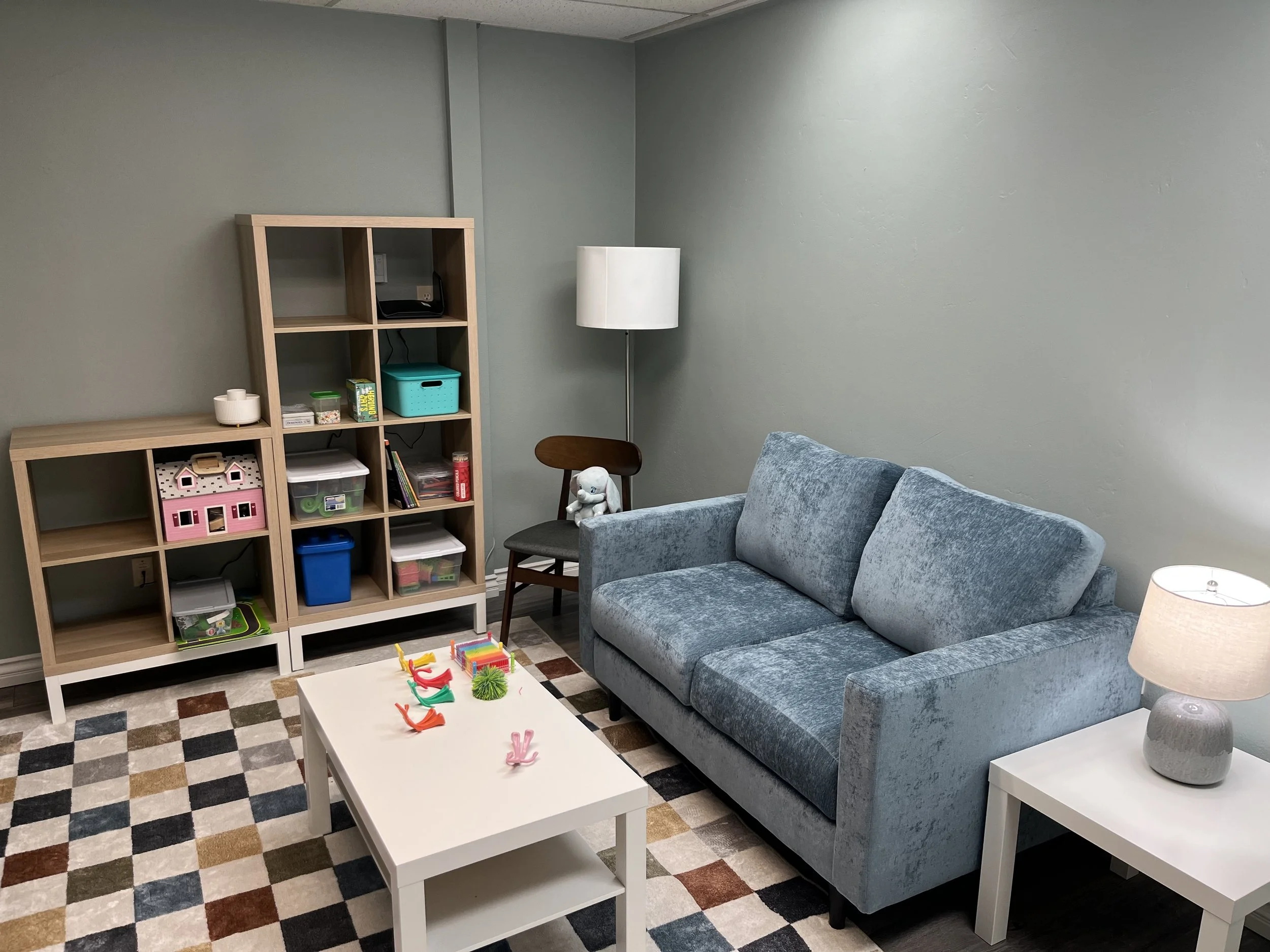 Waiting room has checkered rug, white coffee table with toys scattered on it. Cubed bookshelves are on the back wall. Blue couch is against wall