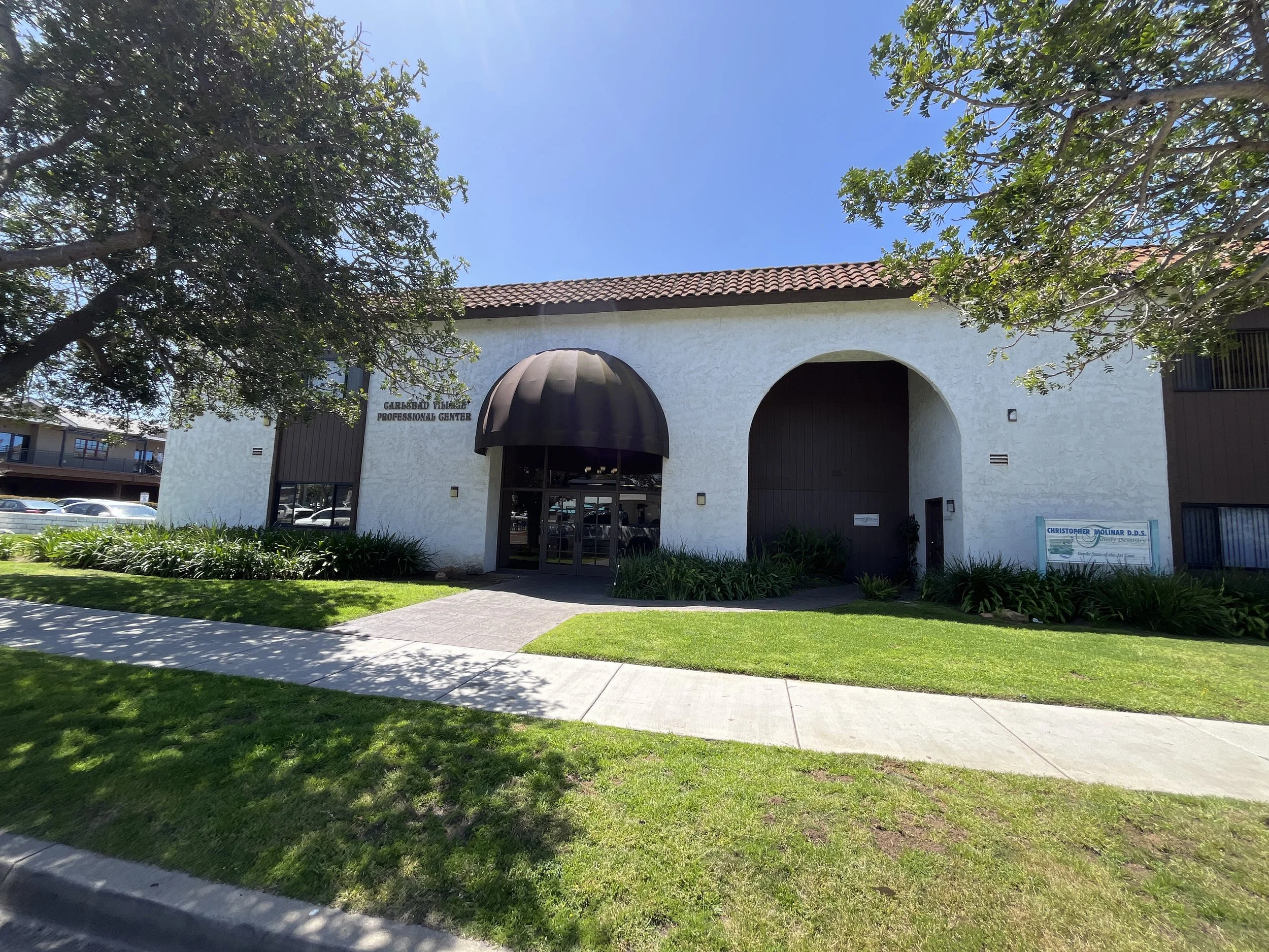 Exterior view of a white building with a red tiled roof, black awning over the entrance, and a sign that reads 'GARDENDALE VILLAGE PROFESSIONAL CENTER'. Green grass, sidewalk, and some trees are in the foreground. Clear blue sky.