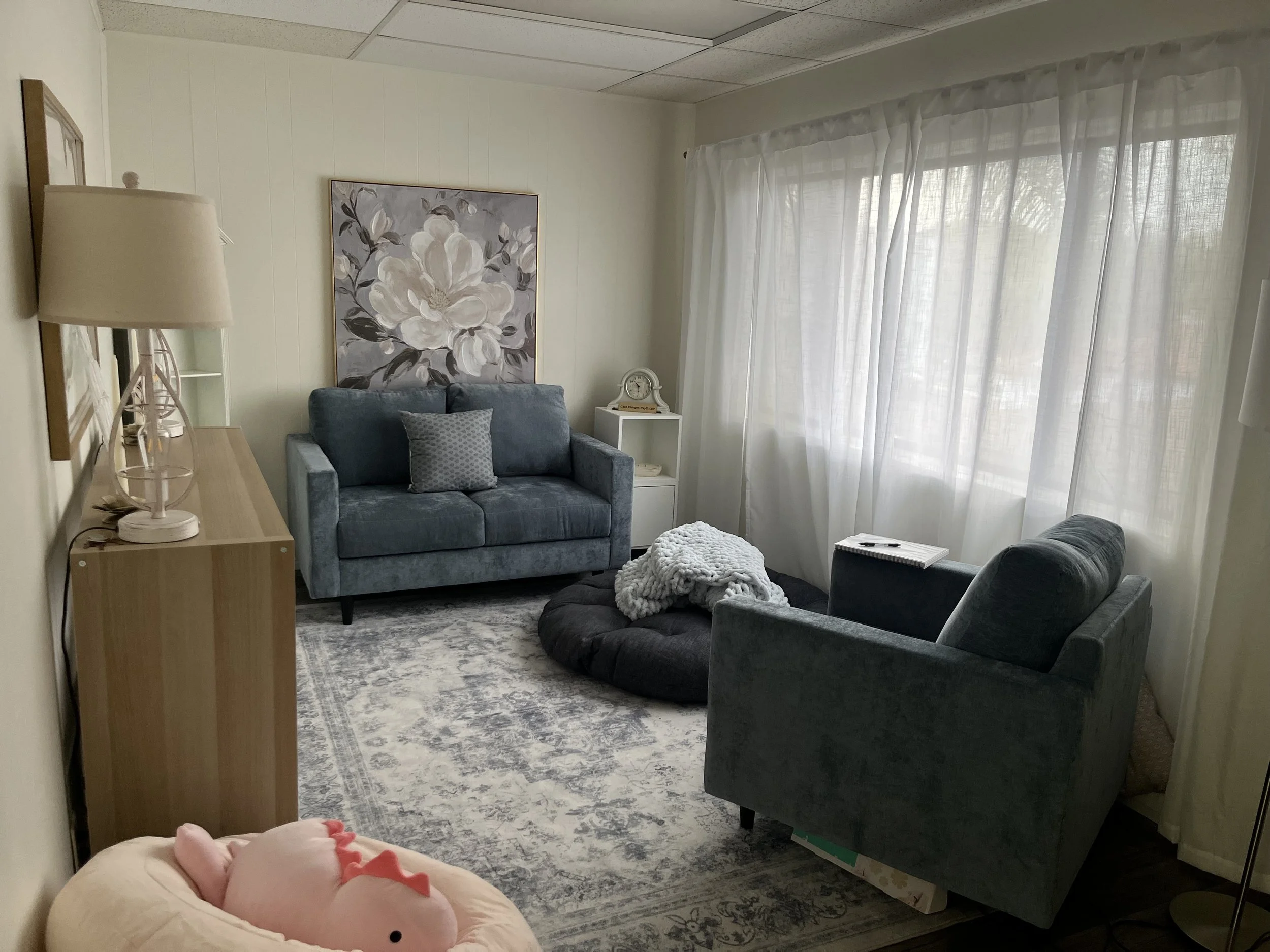 Therapist office with overstuffed chair, facing a couch. Floral print is on the wall. There is a pink stuffed dinosaur on an stuffed bean bag on the fllor