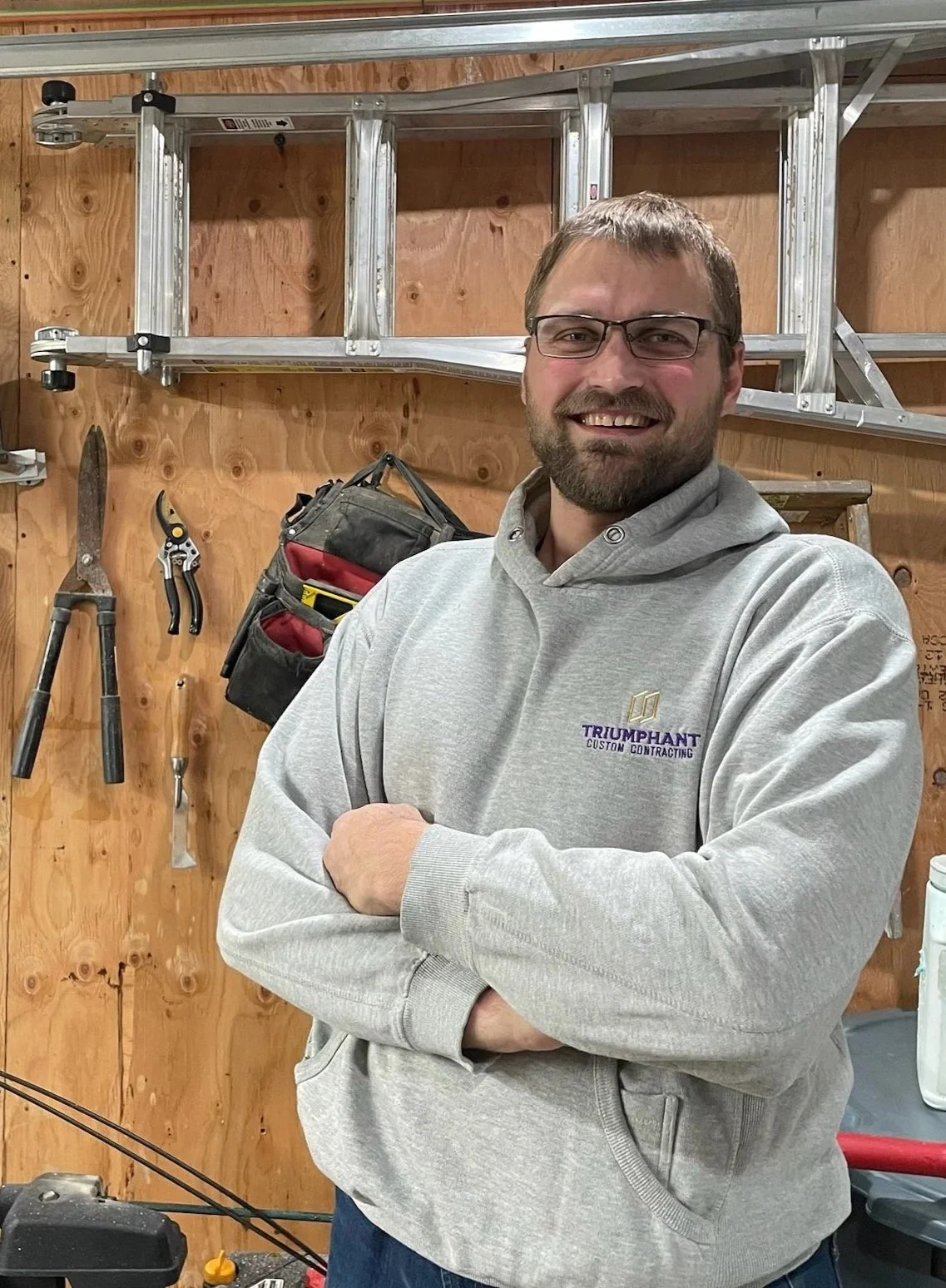 A smiling man with glasses and a beard wearing a gray hoodie with 'Triumphant Custom Contracting' logo, standing in a workshop with tools and a ladder on the wooden wall.