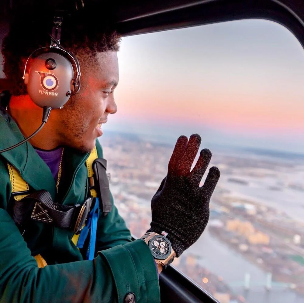 Young man wearing headphones, a watch, and gloves, looking out of helicopter window at cityscape during sunset, waving with hand.