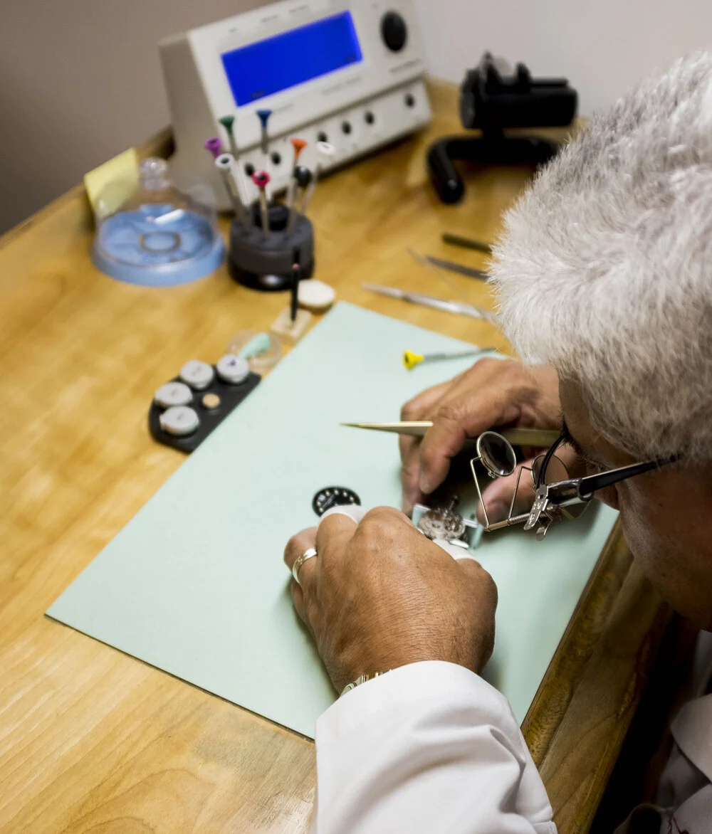 A person working with a watch movement on a green mat, surrounded by watchmaking tools and equipment on a wooden table.