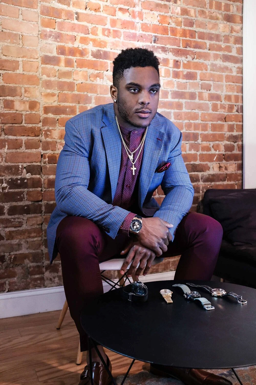 Young man sitting on a chair with a brick wall background, wearing a blue checked blazer, maroon pants, and accessorized with gold chains, a watch, and earrings, with four watches and a camera on the black table in front of him.