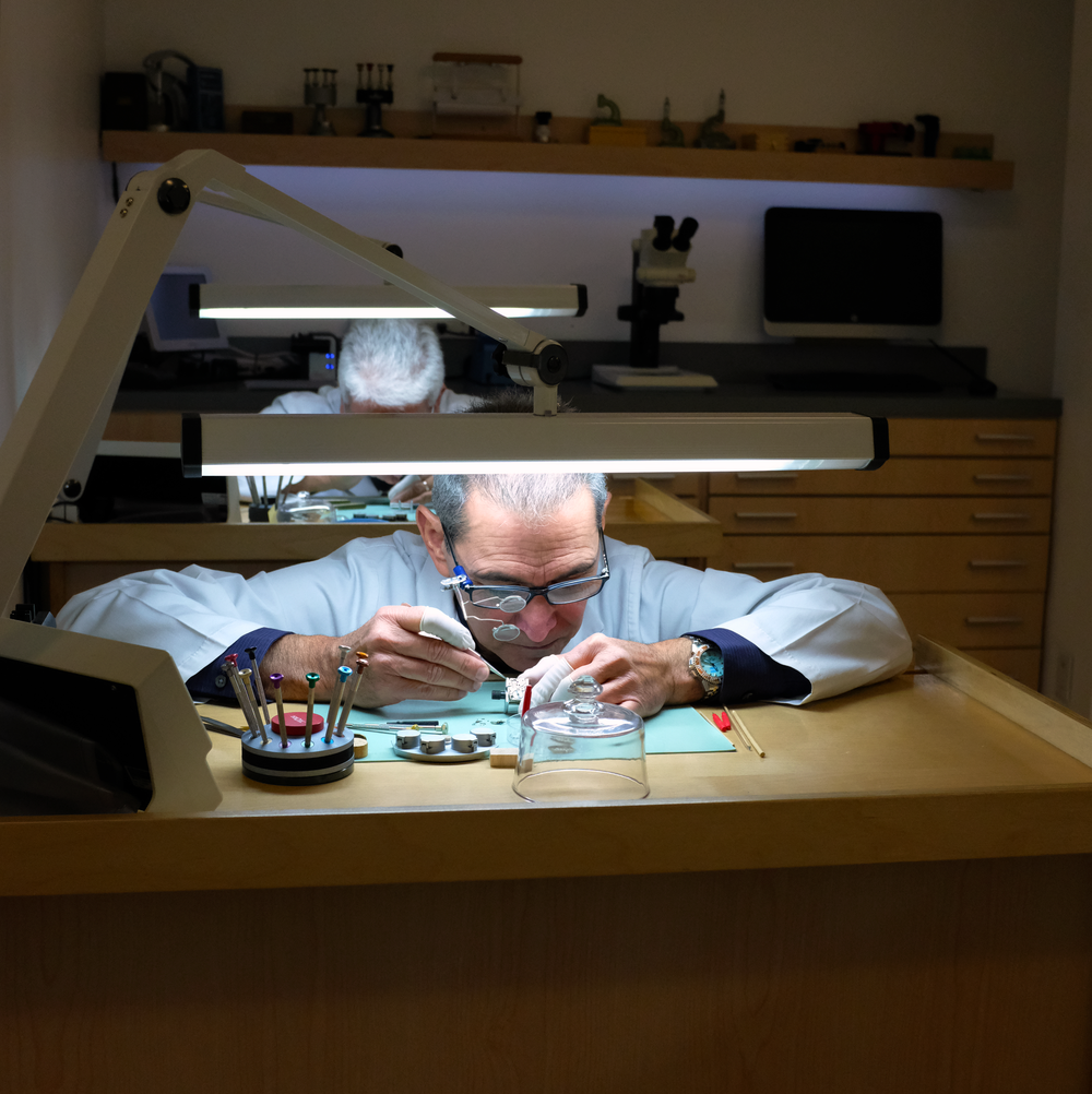 A scientist or engineer working on a small device at a cluttered workbench in a laboratory. The person is wearing glasses, a white coat, and gloves, with a focused expression. The workbench has tools, a glass bell jar, and a circular container with c