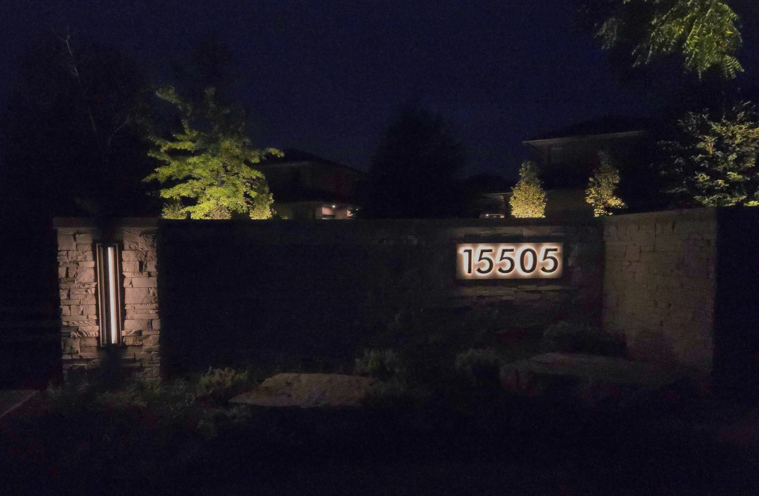 Nighttime view of a house entrance with a stone wall and illuminated house number 15505, surrounded by trees.