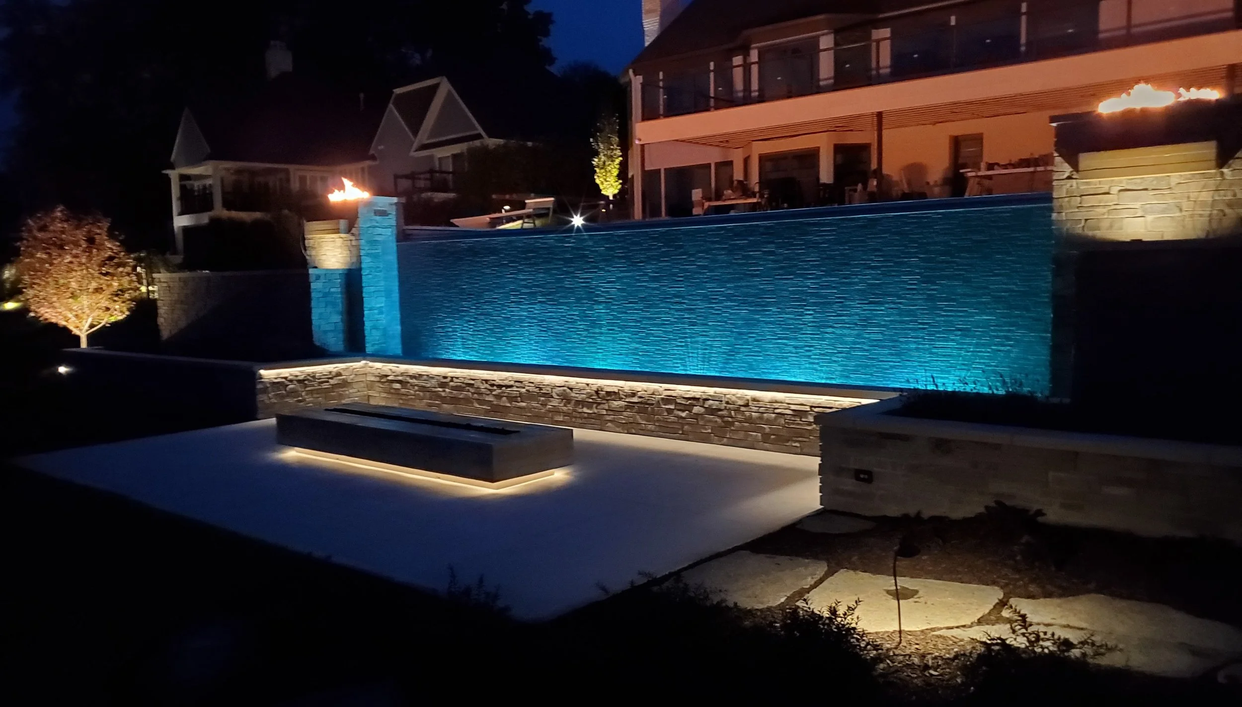 Nighttime view of a backyard with a swimming pool illuminated by underwater lights, a fire feature on a raised platform, and landscaped areas with trees and furniture on an upper patio.