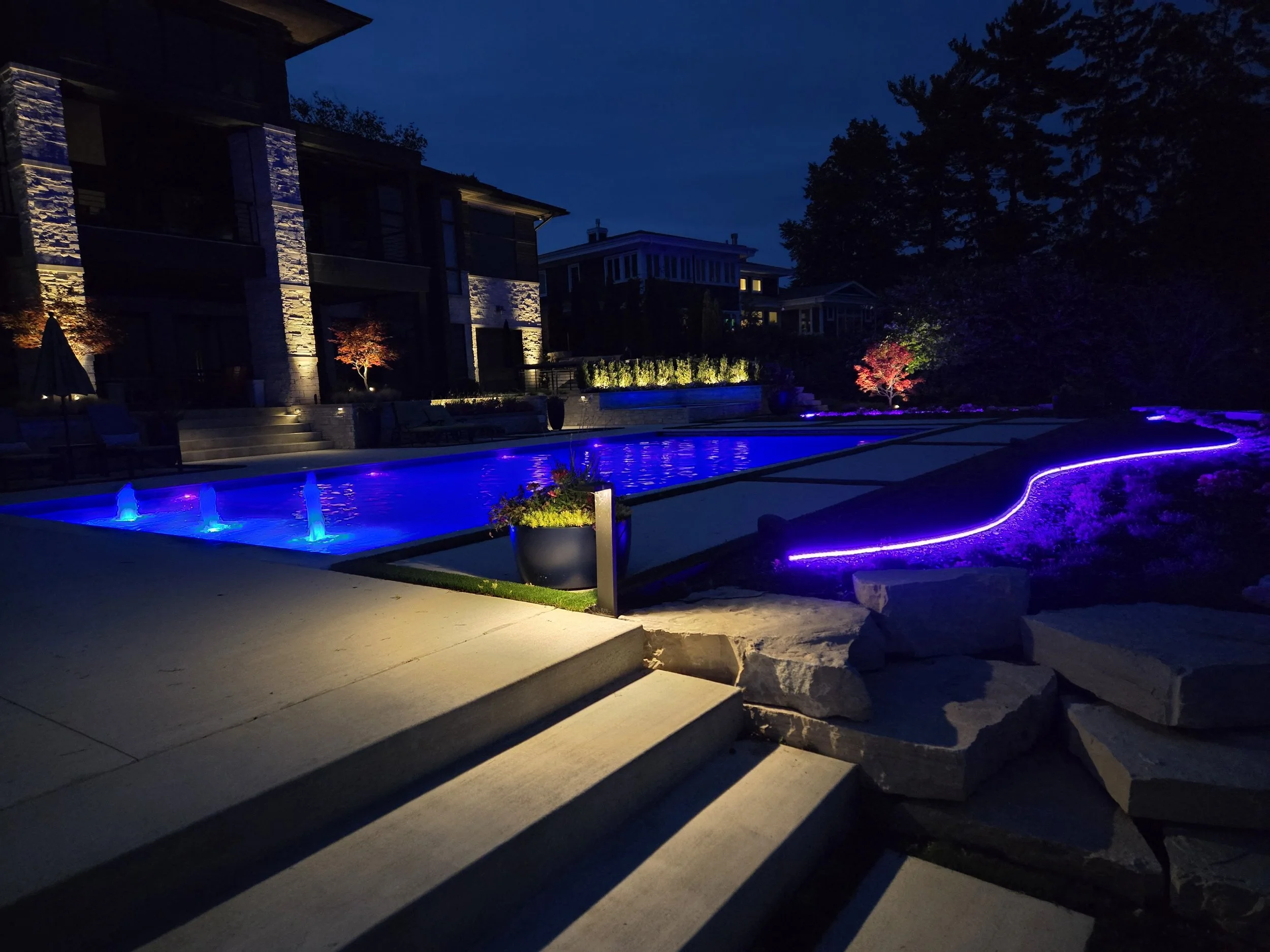 Night view of a modern backyard with a lit pool, illuminated steps, and garden lighting including trees with red and purple lights.