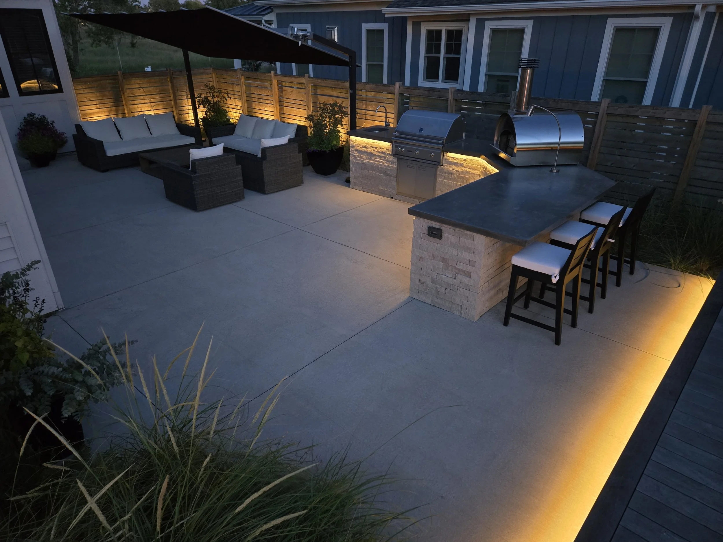 A modern outdoor patio with seating area, grill, and bar, illuminated by warm LED lighting at dusk.