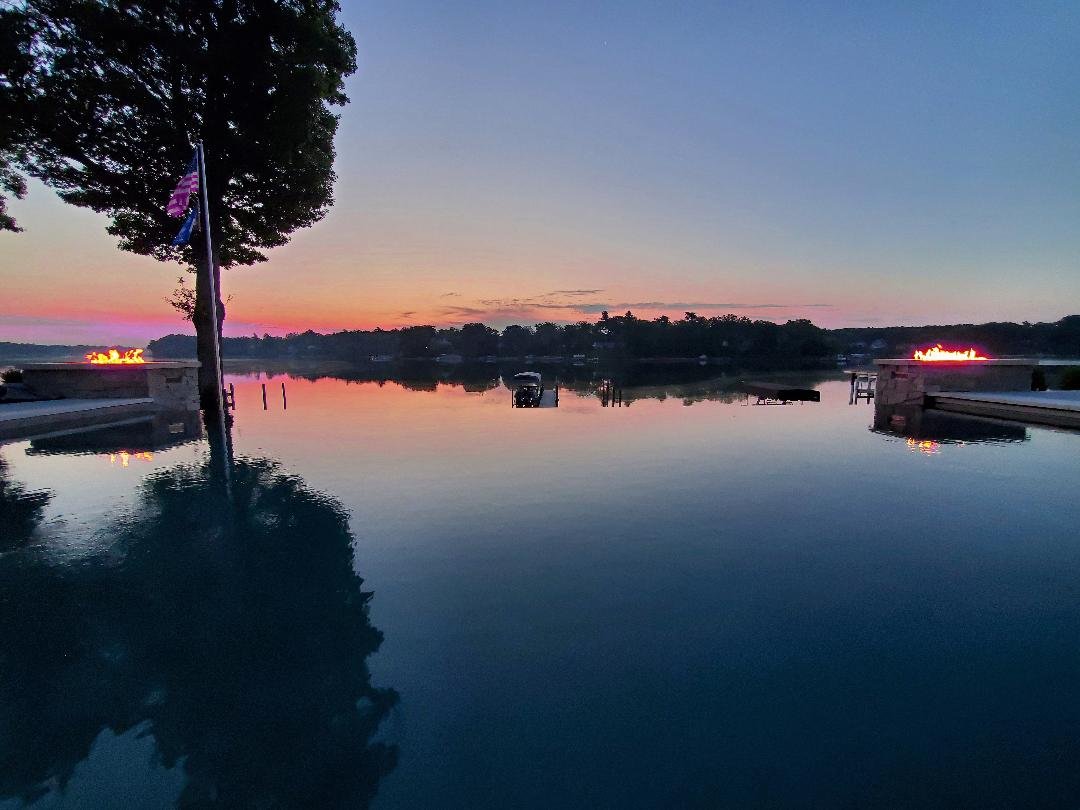 Calm lake or river at sunset with a clear sky, trees and dock on both sides, flags on a tree, and glowing neon signs in the background.