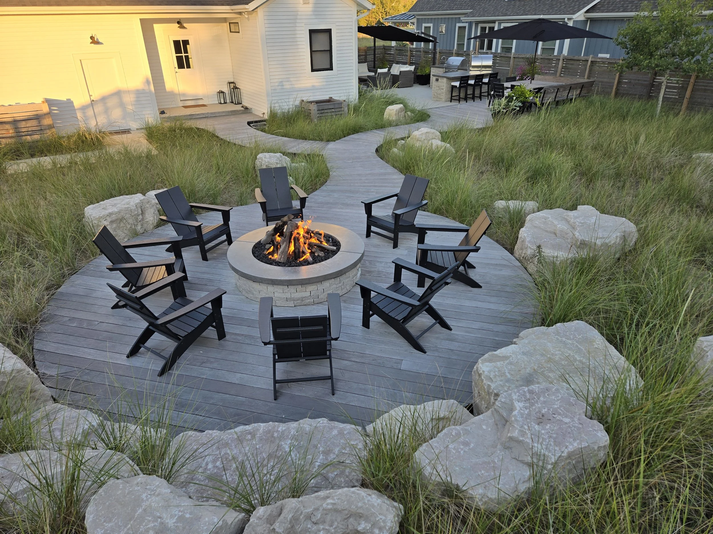Outdoor backyard with a circular fire pit surrounded by black chairs set on a wooden deck, with rocks and greenery around, and a pathway leading to a patio with tables, chairs, and umbrellas.