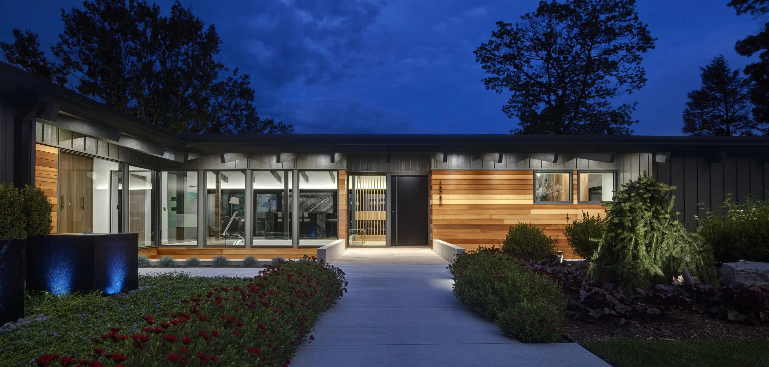 Modern house exterior illuminated at night with wood paneling and large glass windows, surrounded by landscaped garden with shrubs and trees.
