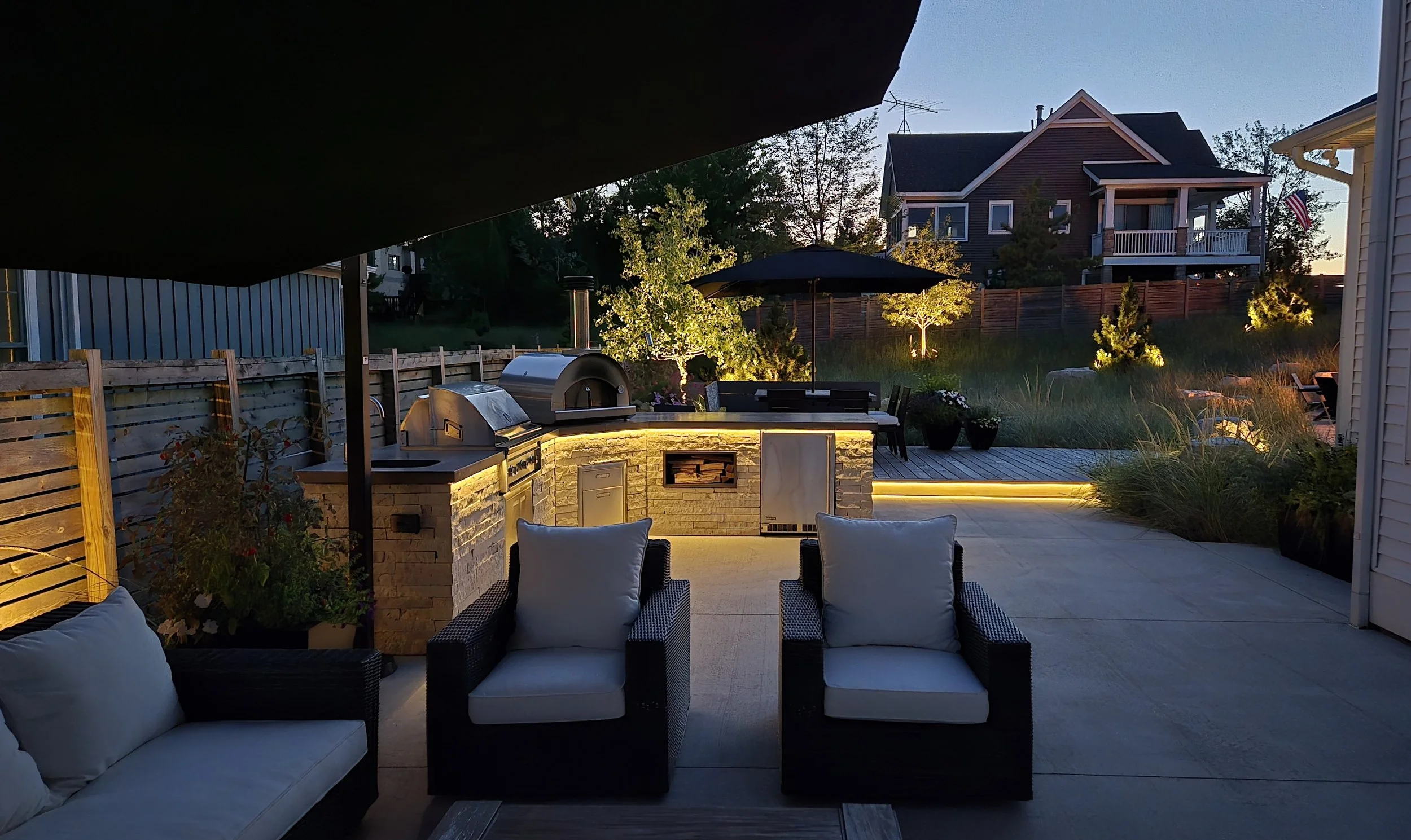 Outdoor patio area with built-in grill and outdoor kitchen, lounge chairs with cushions, potted plants, and landscaped yard with trees and neighboring houses in the background during dusk.