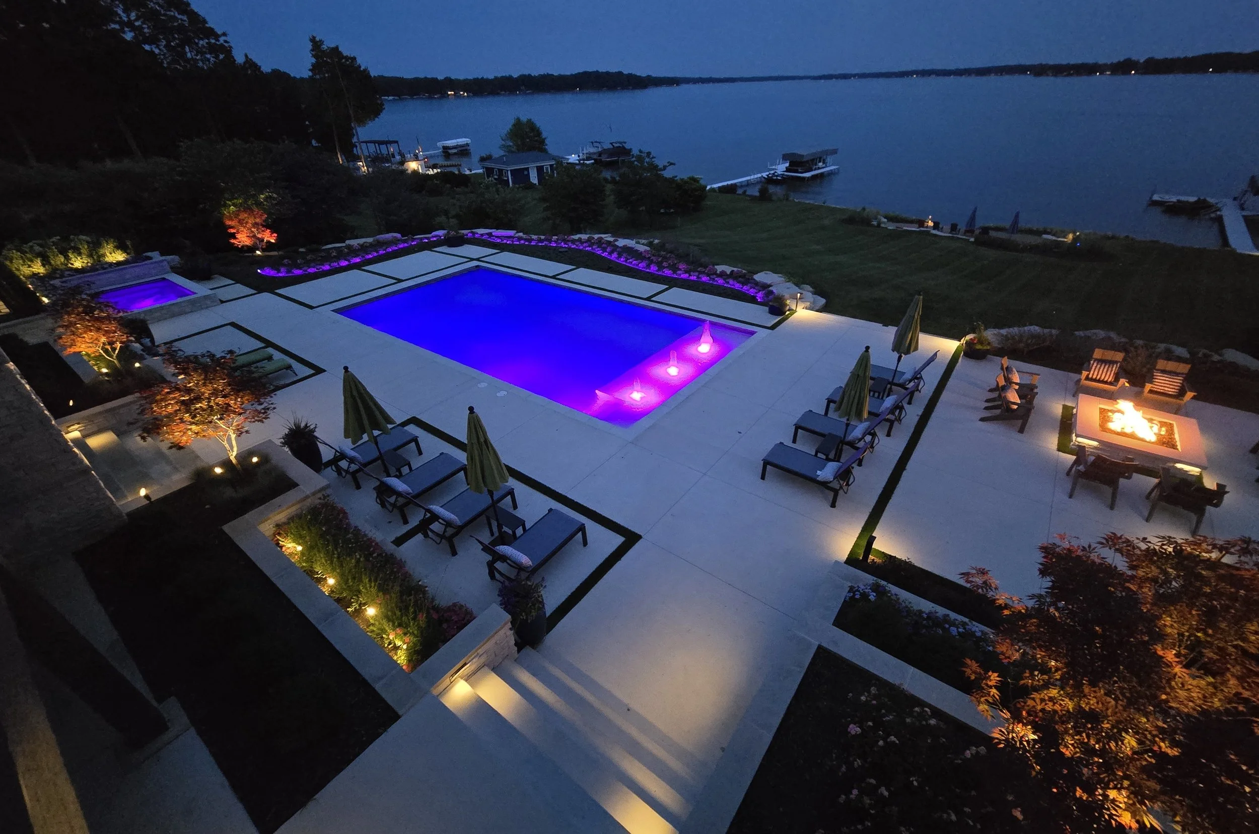 Aerial view of a waterfront backyard at night featuring a lit swimming pool with purple and blue lights, outdoor furniture with umbrellas, a fire pit with chairs, well-lit garden beds, and a view of a lake with boats.