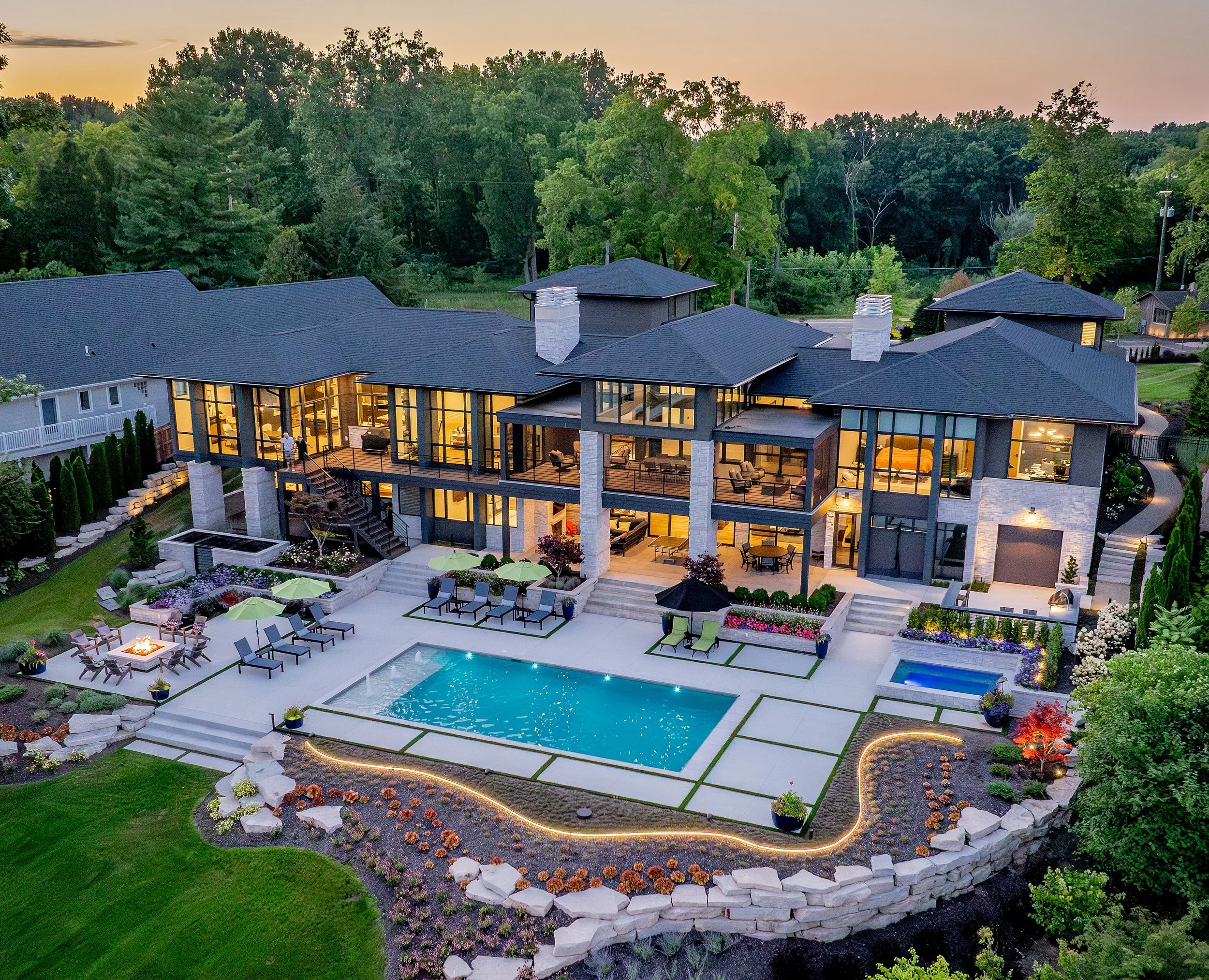 A modern house with large glass windows, view of a backyard with a swimming pool, patio furniture, and landscaped gardens, during dusk.