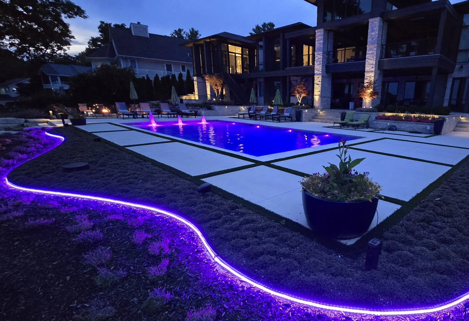 A backyard with a lit-up swimming pool with purple underwater lights and fountains, surrounded by lawn with purple LED lighting along the edge, chairs, and potted plants, with a large house in the background.