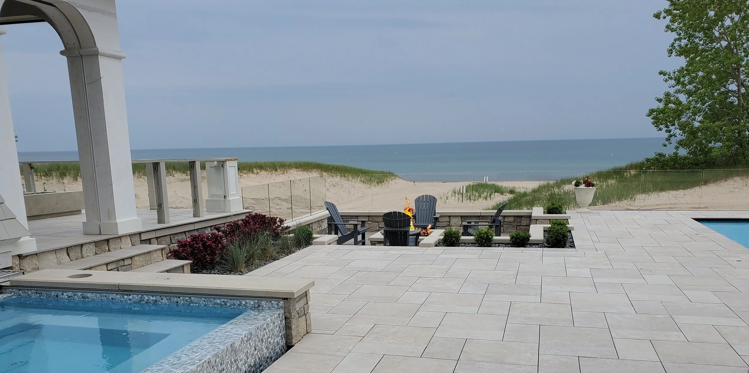 Patio area with four black Adirondack chairs surrounding a fire pit, overlooking a sandy beach and ocean in the background, with a swimming pool on the right and a large potted plant on the right side.