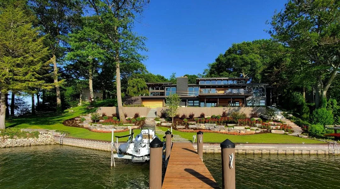 Modern waterfront house with large windows, landscaped yard, and dock with boat, set among tall trees under a clear blue sky.