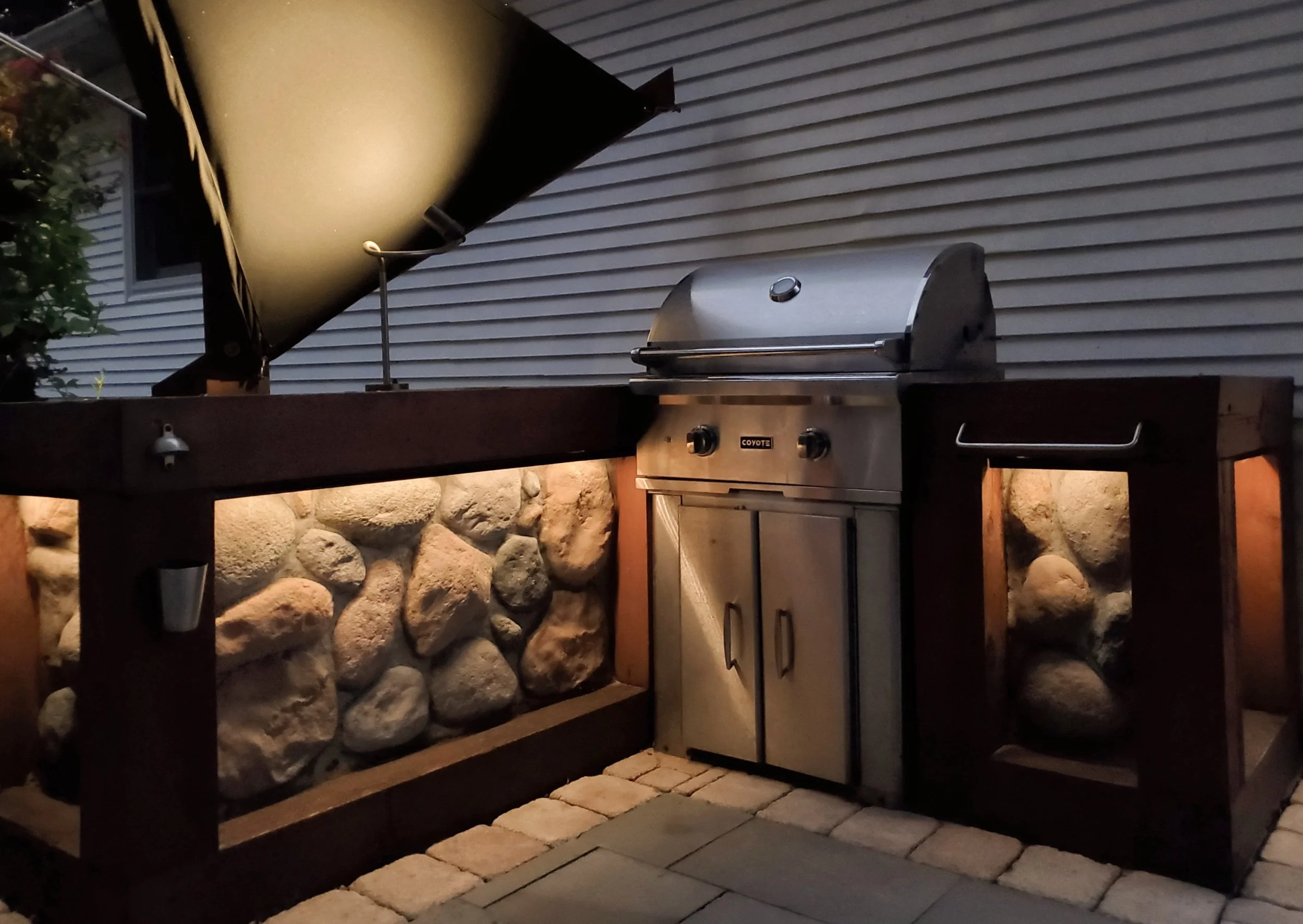 Outdoor kitchen area with a built-in stainless steel gas grill, a stone wall feature, and a large black shade sail overhead, set against a house with white siding.