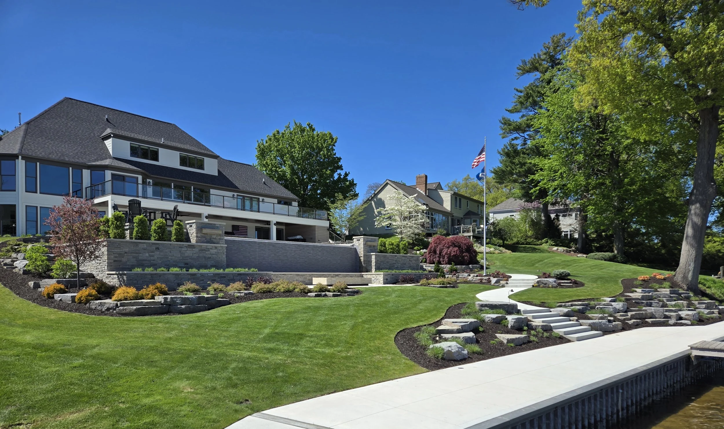 Luxury house with a large terrace, manicured lawn, colorful plants, trees, and a flagpole under a clear blue sky.