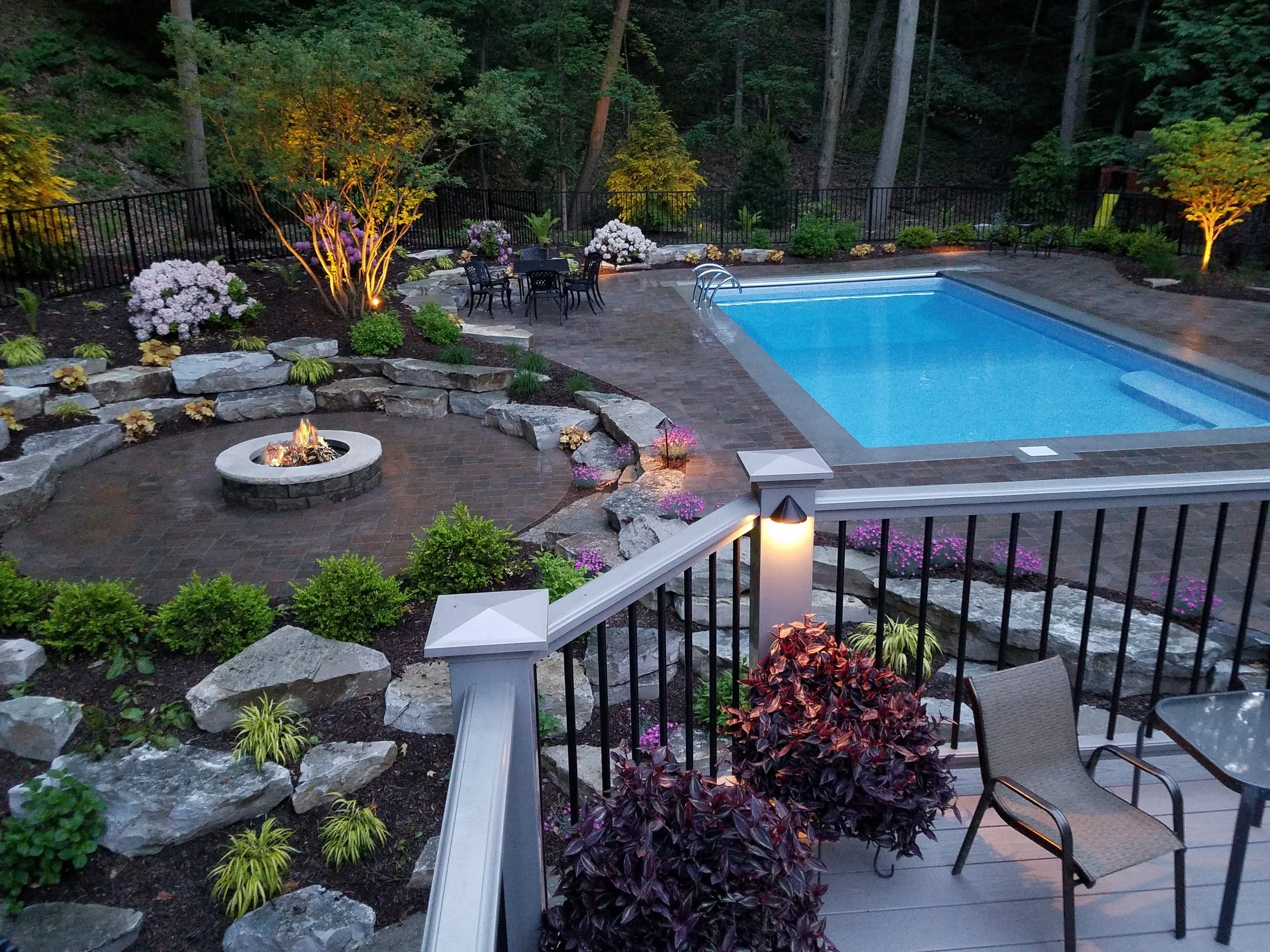 Backyard with a swimming pool, stone fire pit, outdoor table with chairs, landscaped garden with trees and shrubs.