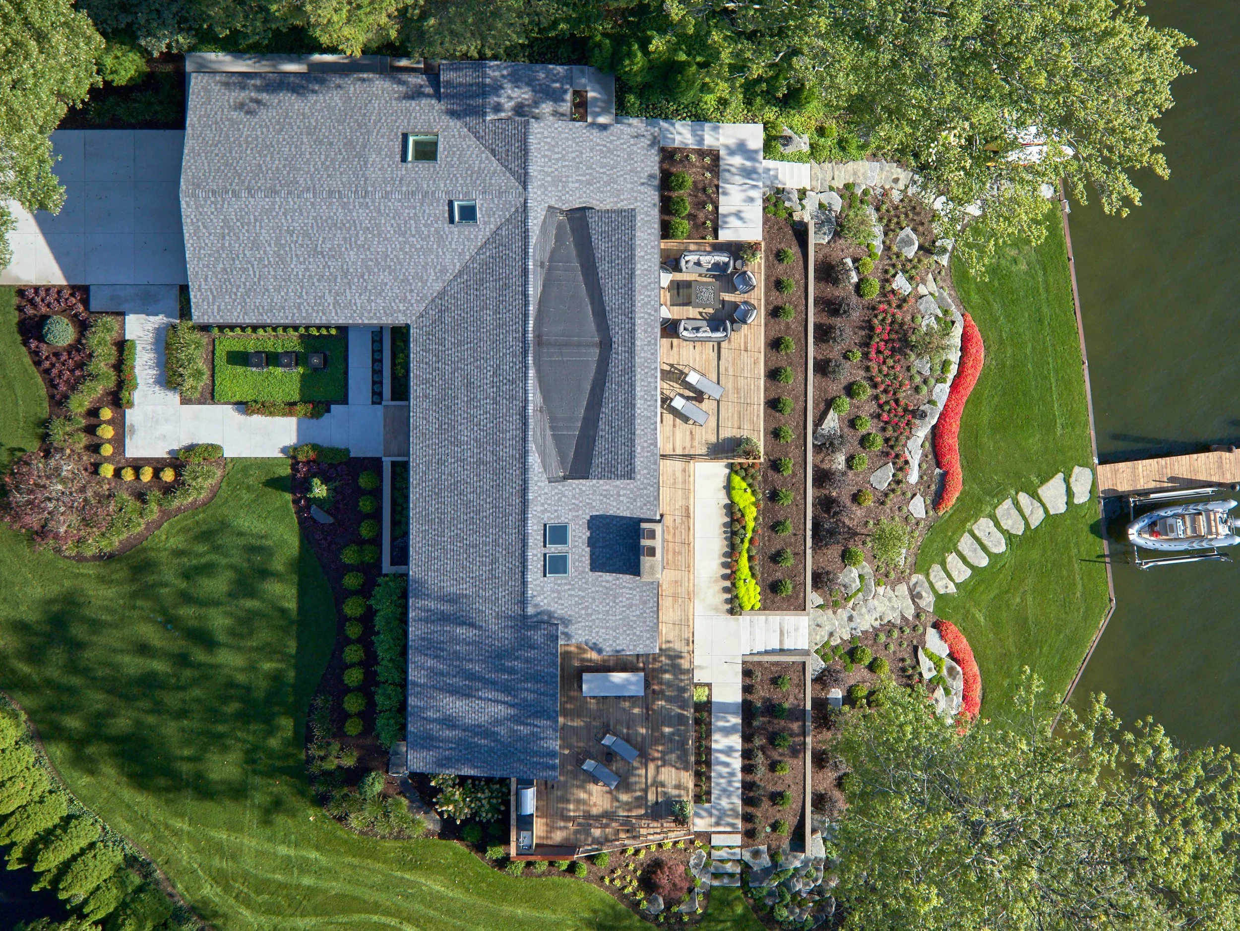Aerial view of a house with a gray roof, landscaped garden, wooden deck with outdoor furniture, and a boat dock by a water body.